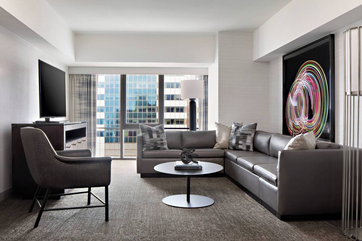 Een moderne hotelkamer met een grijze bank, een ronde salontafel, een flatscreen televisie en een groot abstract schilderij.