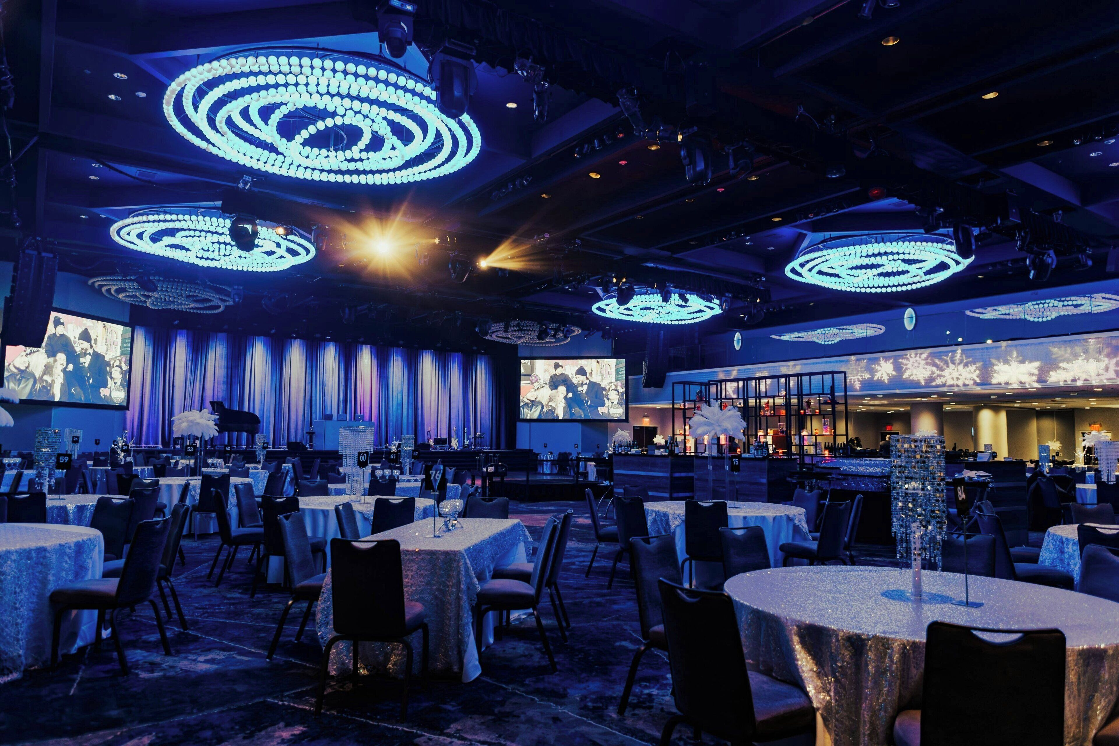 A spacious banquet hall with round tables, a stage with curtains, and a large screen displaying an image.