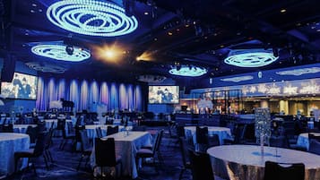 Un amplio salón de banquetes con mesas redondas, un escenario con cortinas y una gran pantalla mostrando una imagen.