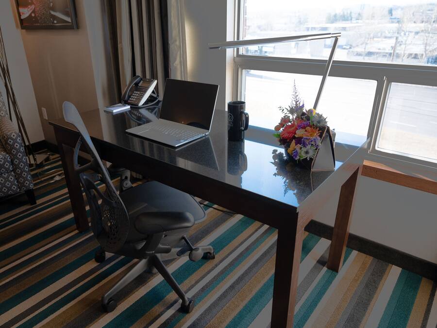 A hotel room with a desk, laptop, phone, mug, and a flower vase.