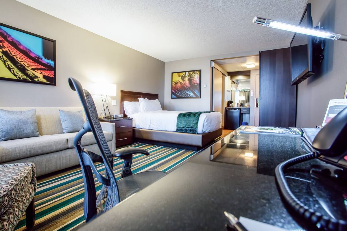 A hotel room with a bed, a desk with a computer, a chair, a sofa, a TV, and a painting on the wall.