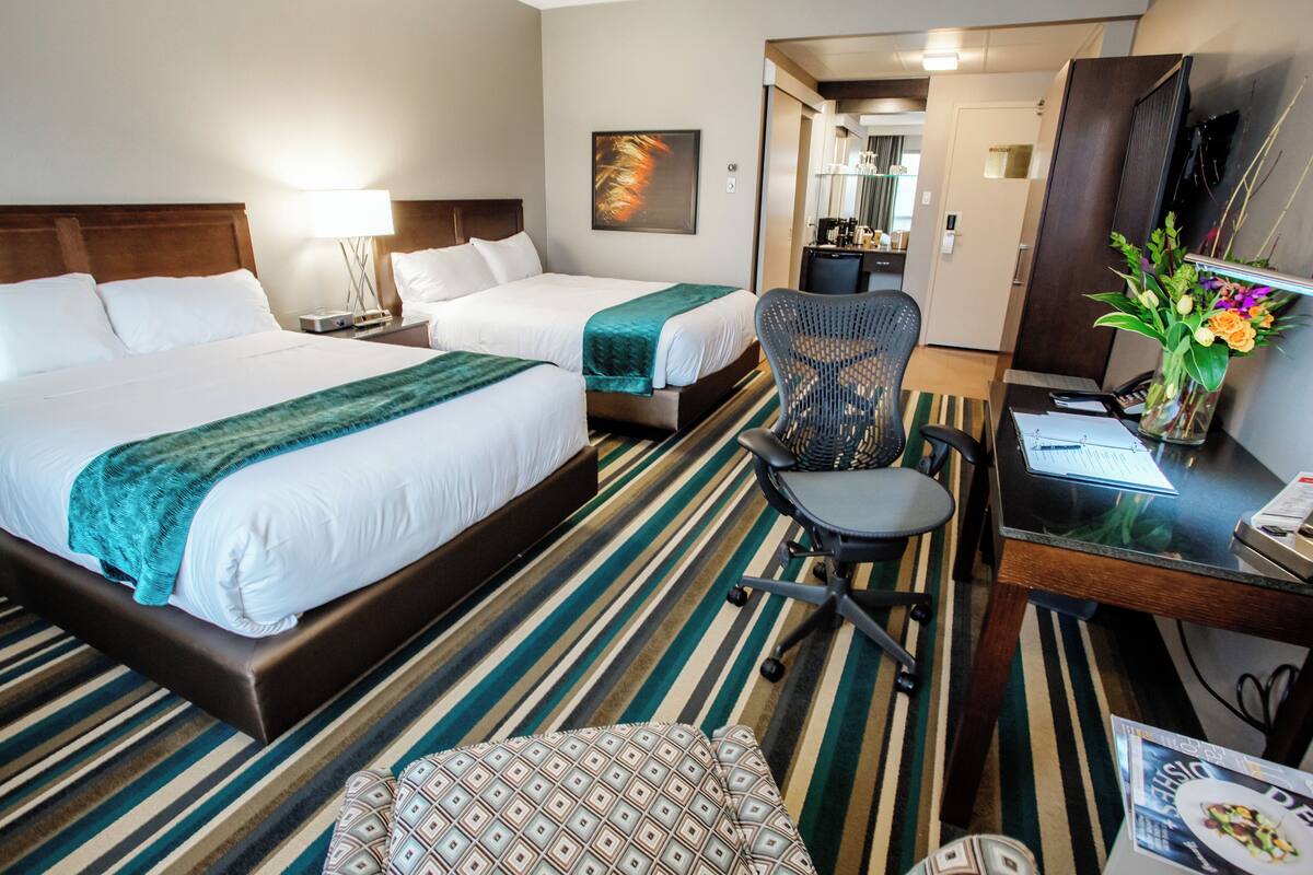 A hotel room with two beds, a desk with a chair, a small table, a lamp, and a decorative vase with flowers.