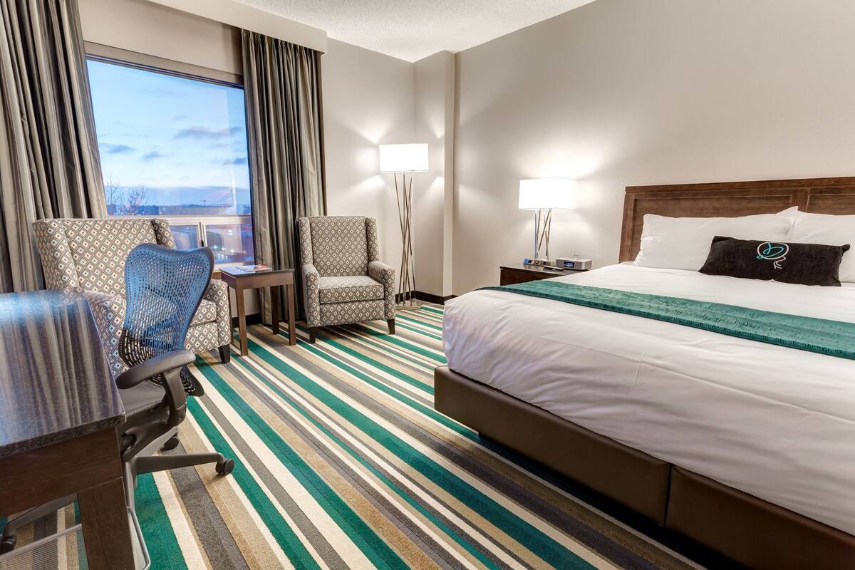 A hotel room with a bed, a desk, a chair, a floor lamp, and a view of the outdoors.