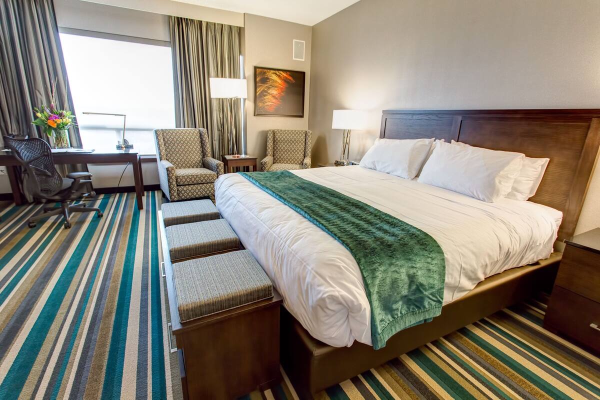 A hotel room with a large bed, two armchairs, a desk with a chair, a lamp, and a window with curtains.