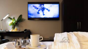 A neatly made bed with a white bathrobe, a tray with a cup and a pitcher, and a television displaying a blue image in the background.