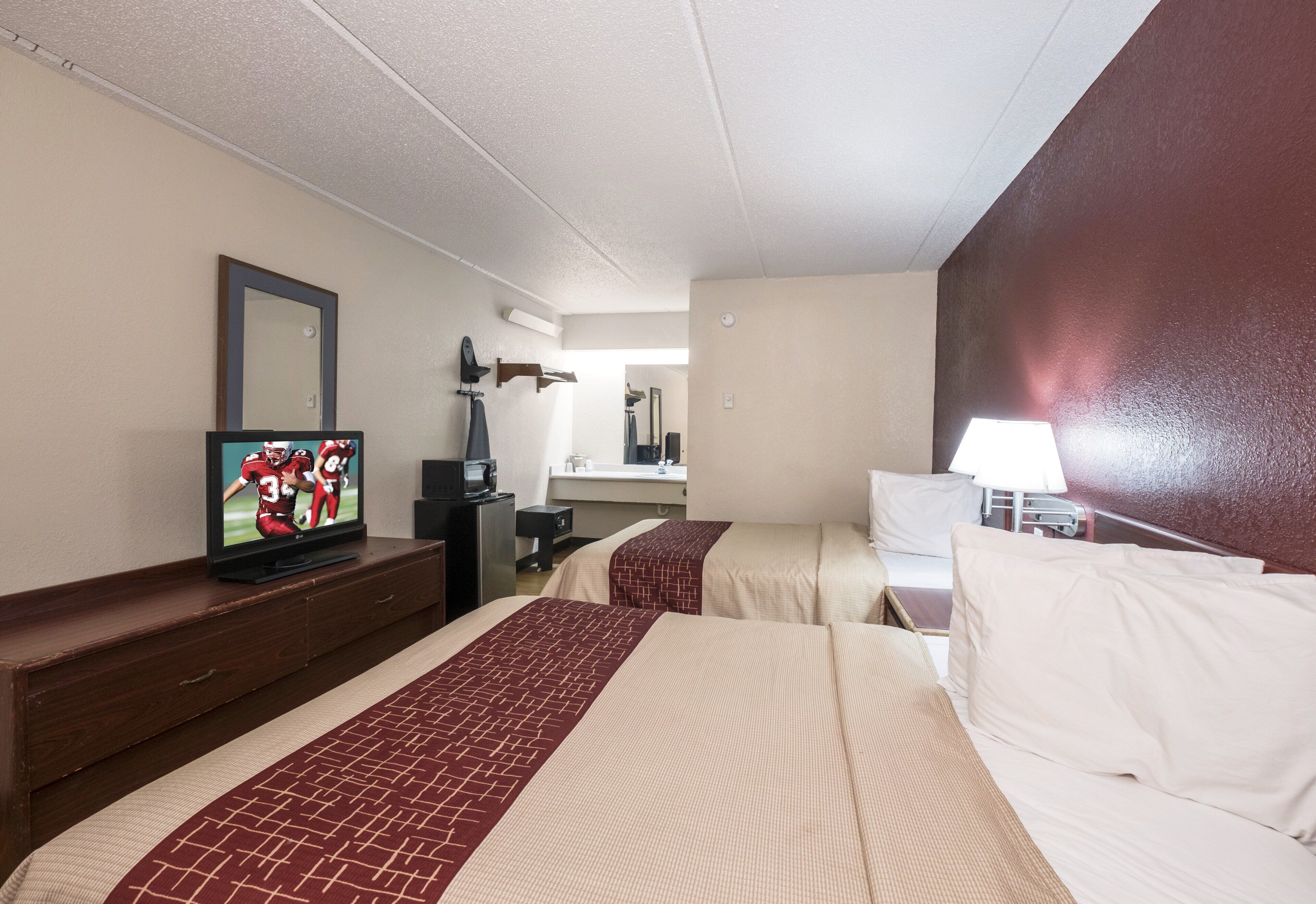 A hotel room with two beds, a TV on a dresser, a desk with a lamp, and a mirror on the wall.