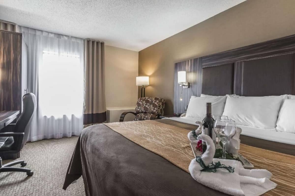 A hotel room with a bed, a desk, a chair, a lamp, and a table with a towel and wine glasses.