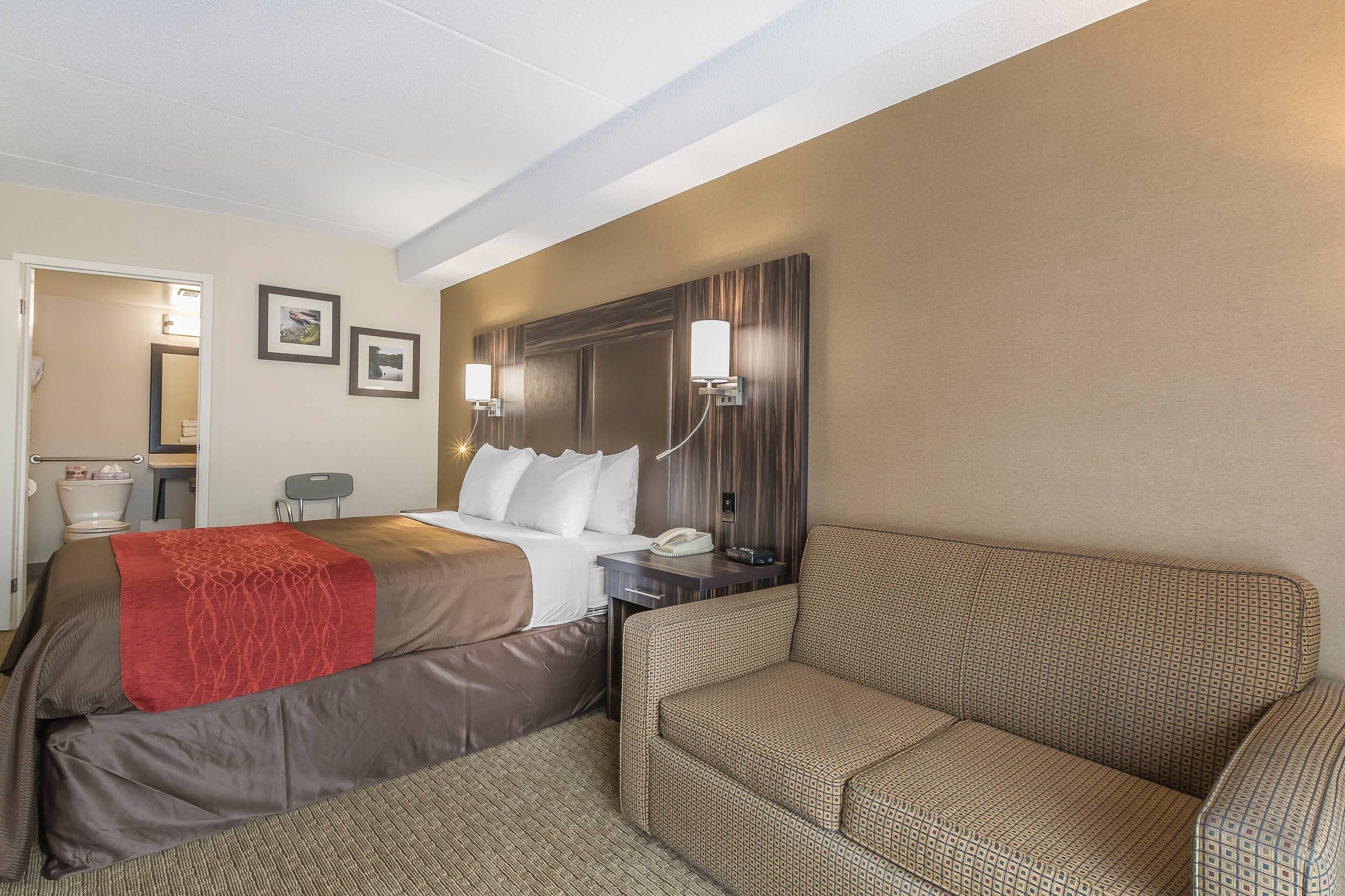 A hotel room with a bed, a sofa, a nightstand with a phone, and a wall-mounted light fixture.