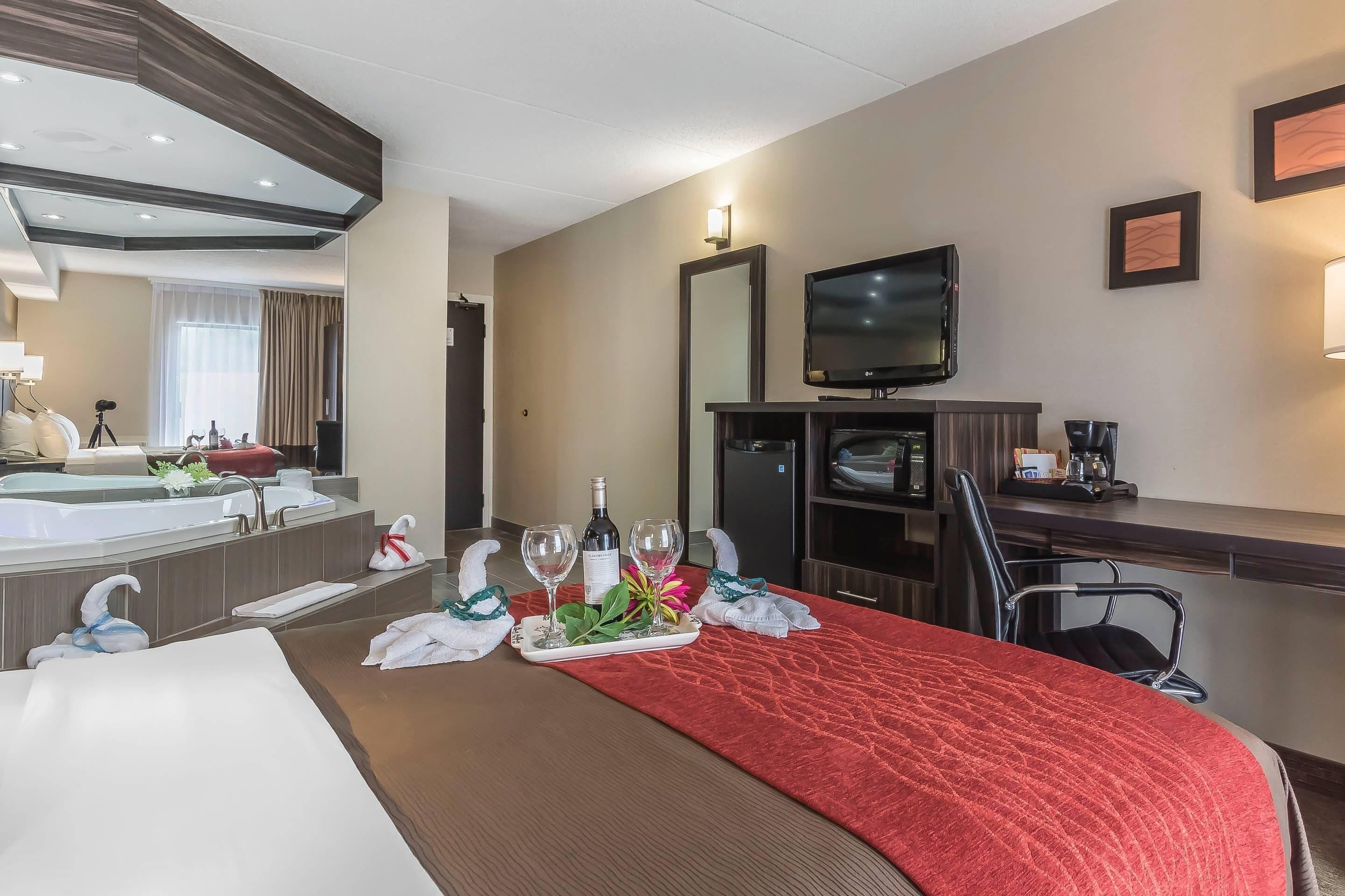 A hotel room with a red carpet, a large bed, a TV, a minibar, a desk, and a bathroom with a bathtub and a mirror.