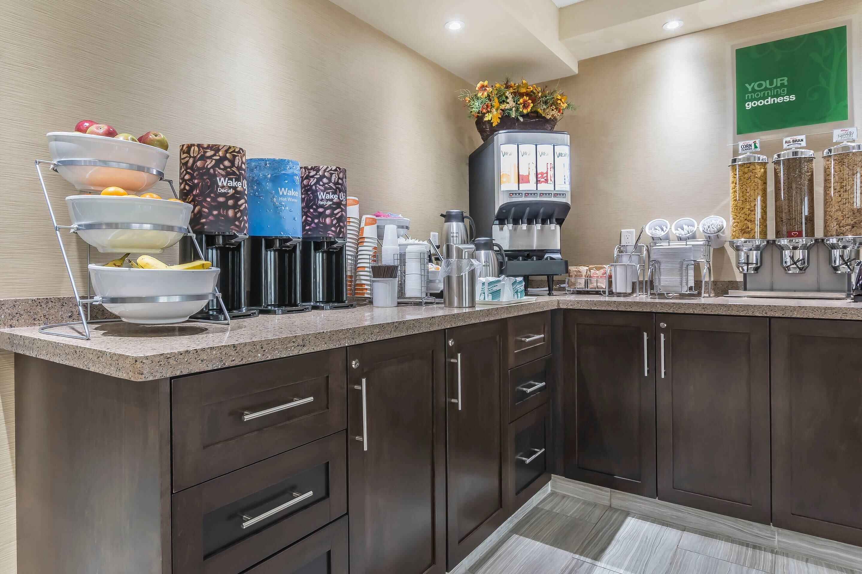 Un espace pour le petit-déjeuner à l'hôtel, avec un buffet où les clients peuvent se servir eux-mêmes, comprenant des machines à café, un panier de fruits et un distributeur de café.
