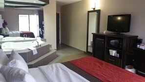 Quarto de hotel com cama, televisão, banheiro com banheira, espelho e escrivaninha com cafeteira.