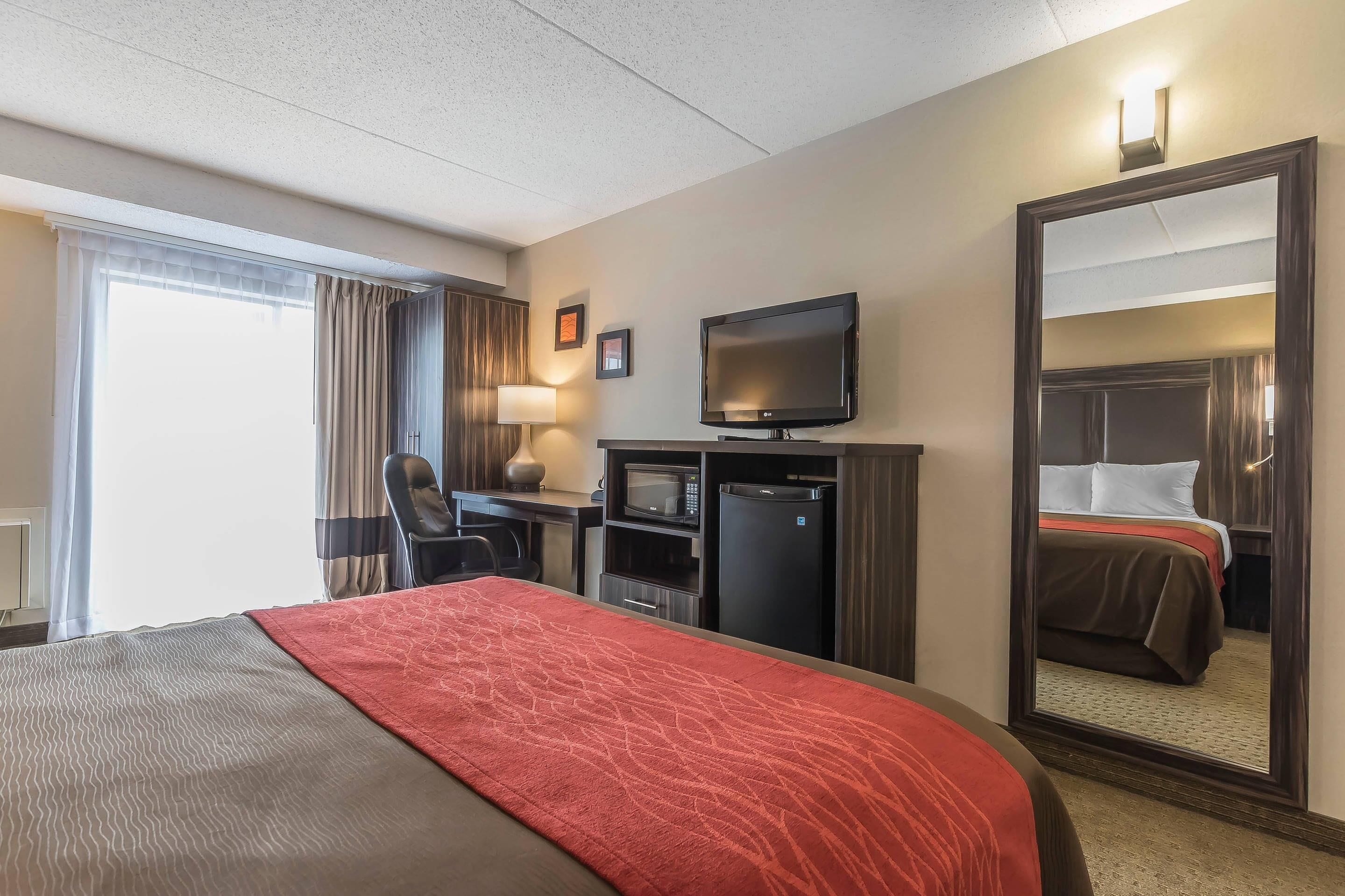 A hotel room with a bed, a desk, a television, a refrigerator, and a large mirror.