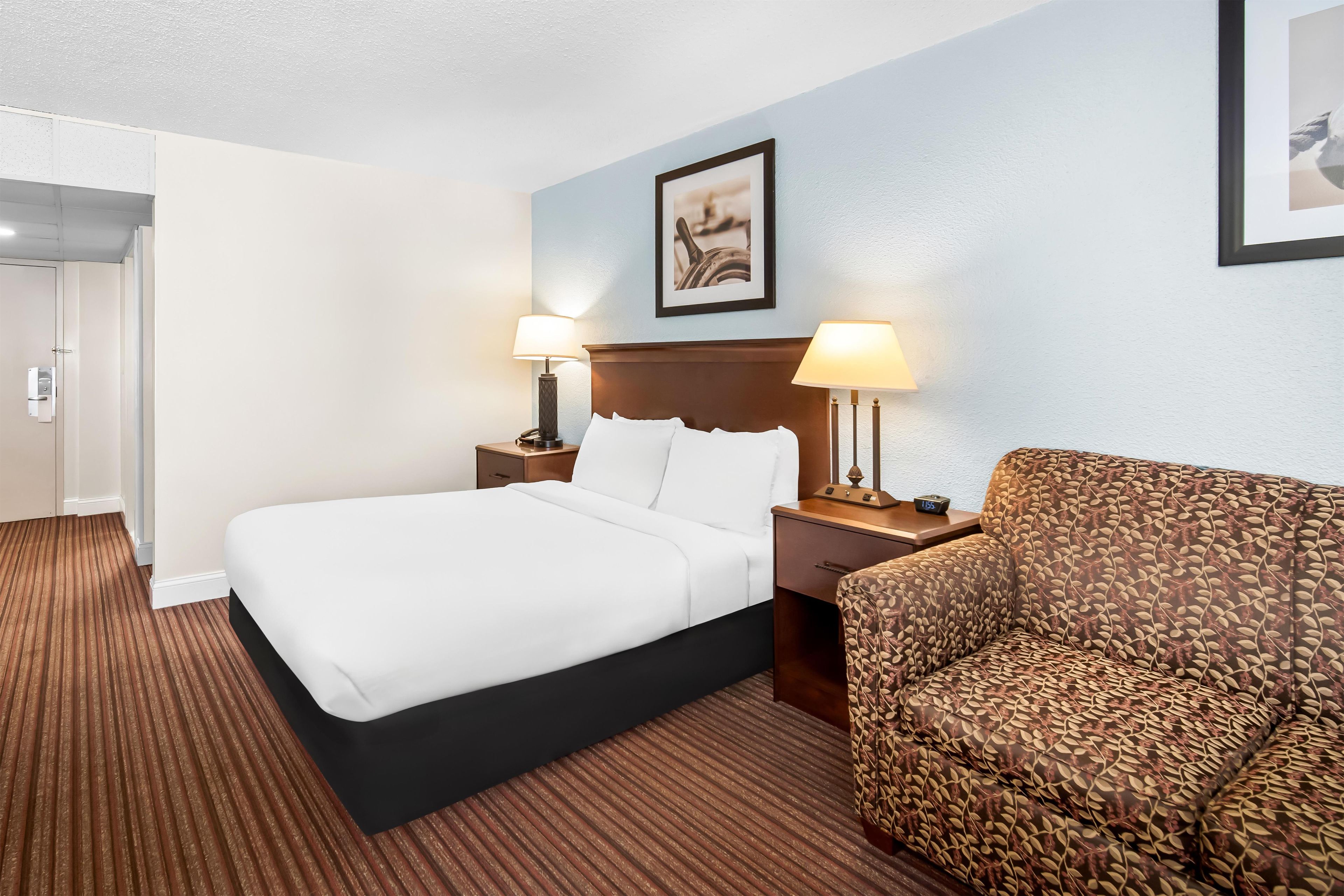 Habitación de hotel con una cama, dos lámparas de noche, una silla, una mesita de noche y un cuadro en la pared.
