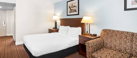 Quarto de hotel com cama, duas luminárias, uma cadeira, mesa de cabeceira e um quadro na parede.