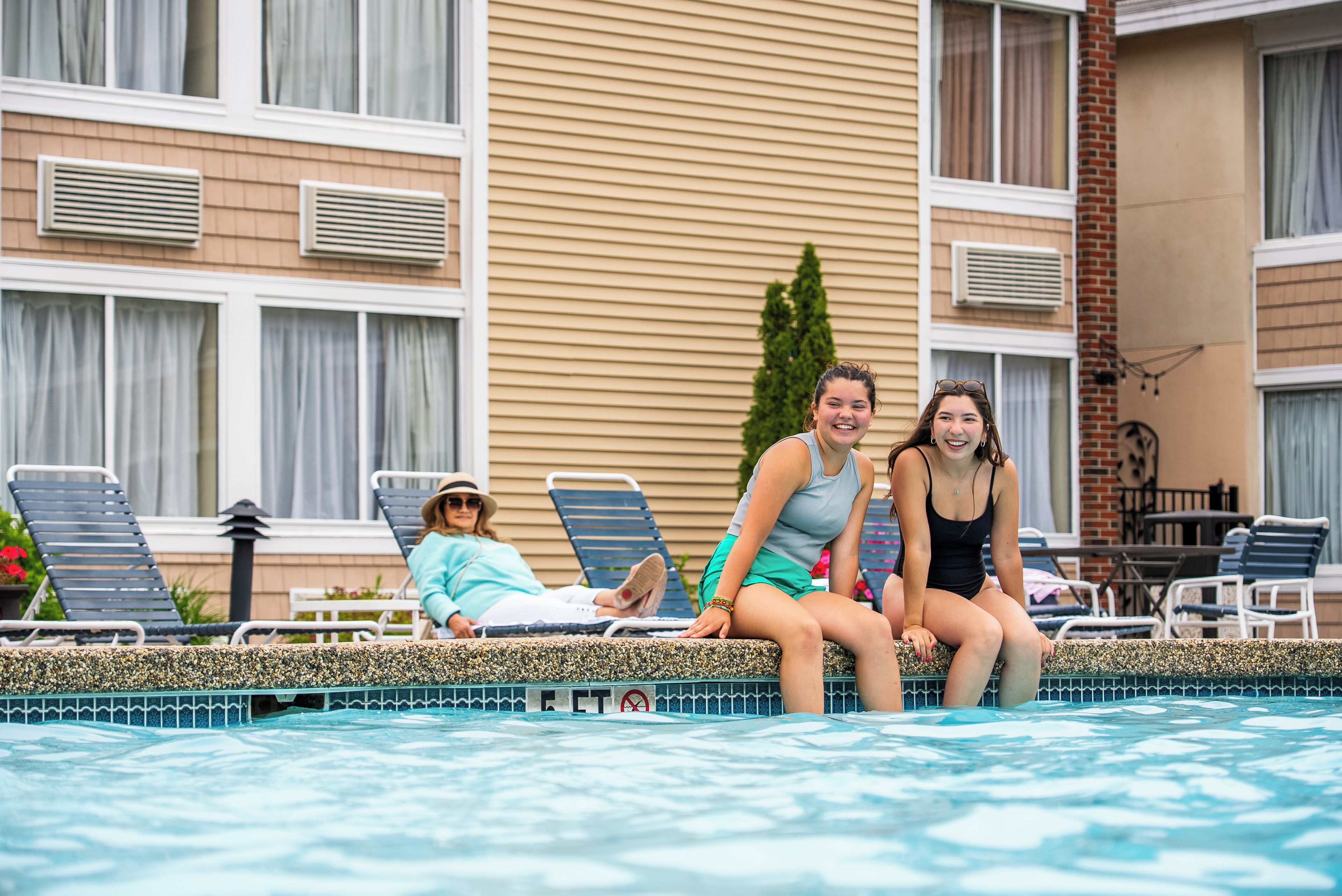 Et poolområde med liggestole og en bygning med mange vinduer samt aircondition i baggrunden.