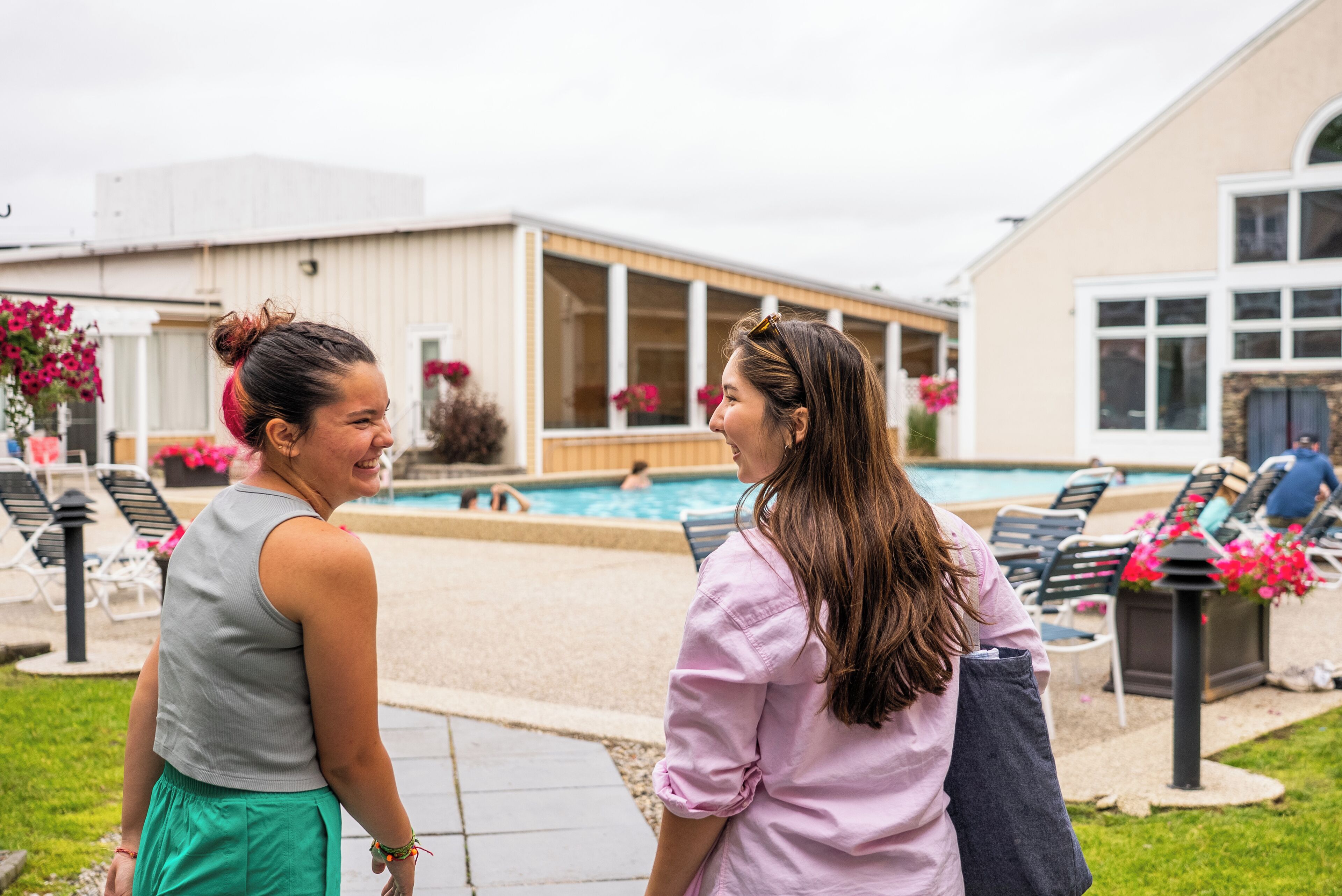 Duas pessoas perto de uma piscina, com espreguiçadeiras e um edifício ao fundo.