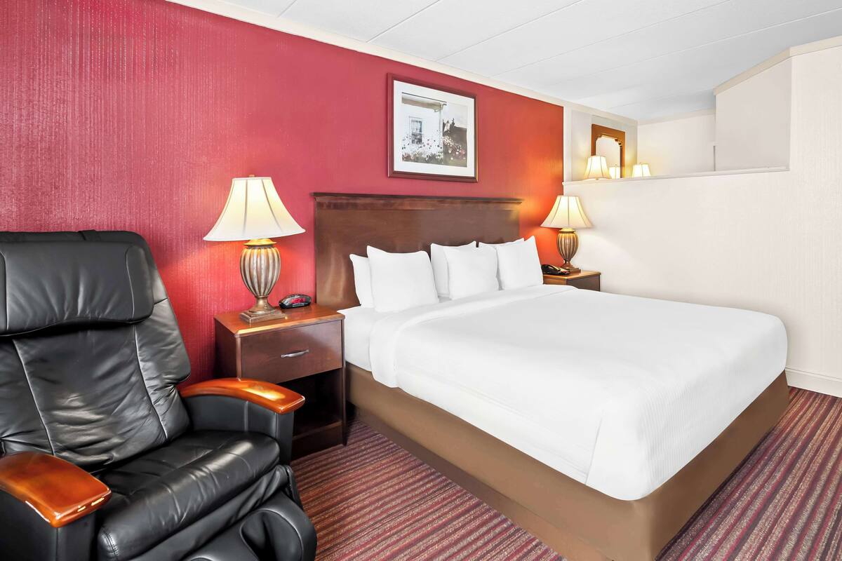 A hotel room with a large bed, a leather armchair, two bedside tables with lamps, and a red accent wall.