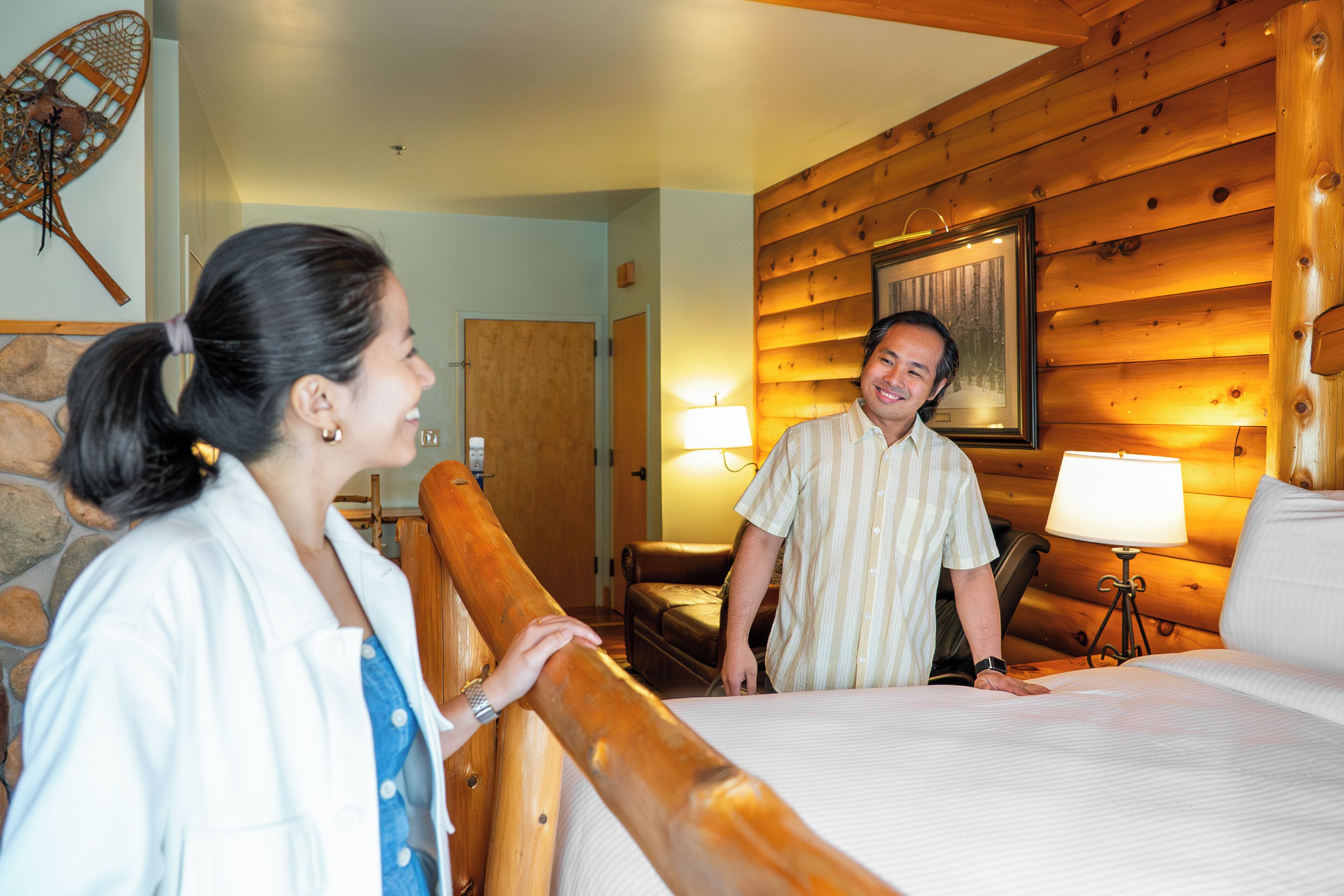 A wooden cabin interior with a bed, a wooden headboard, a wall-mounted lamp, and a framed picture.
