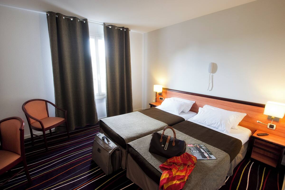 Una habitación de hotel con cama, dos sillas, una mesita de noche, una lámpara, teléfono y ventana con cortinas.