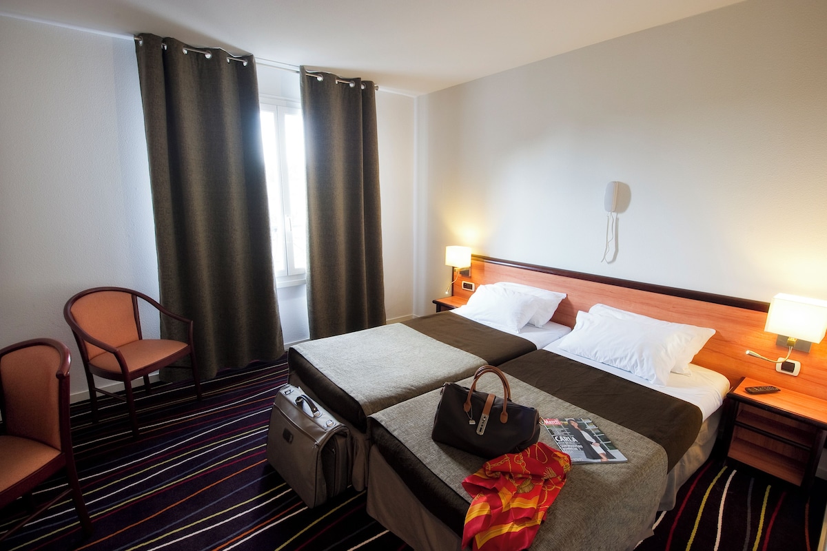 Una habitación de hotel con cama, dos sillas, una mesita de noche, una lámpara, teléfono y ventana con cortinas.
