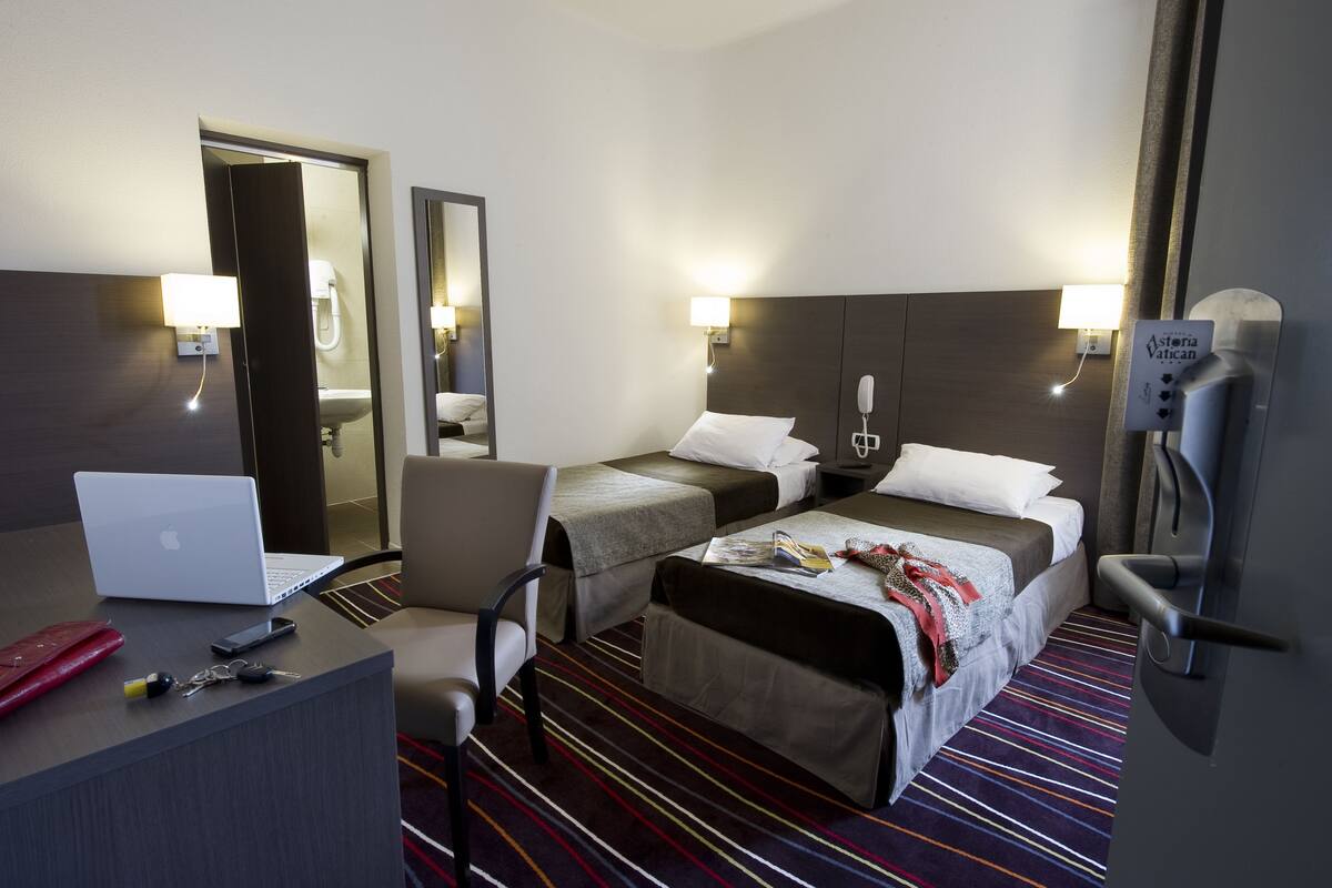 Habitación de hotel con dos camas, un escritorio con una computadora portátil, una silla y un baño visible a través de una puerta abierta.