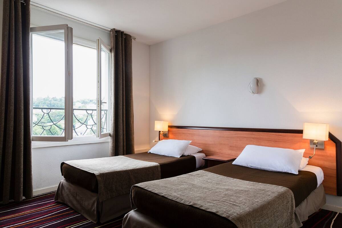 Habitación de hotel con dos camas, una ventana con cortinas, vista a zonas verdes y una lámpara fijada a la pared.