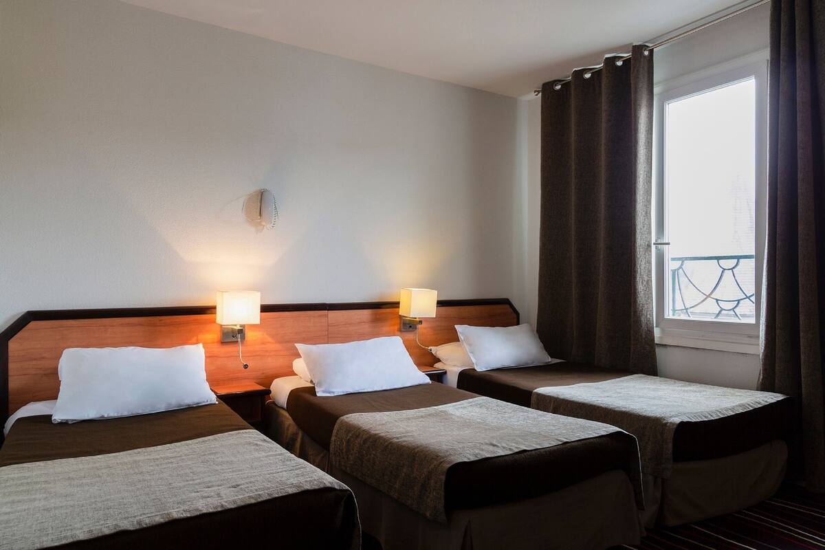 Habitación de hotel con tres camas individuales, cada una con almohada y manta. Dos lámparas de pared a ambos lados de las camas.