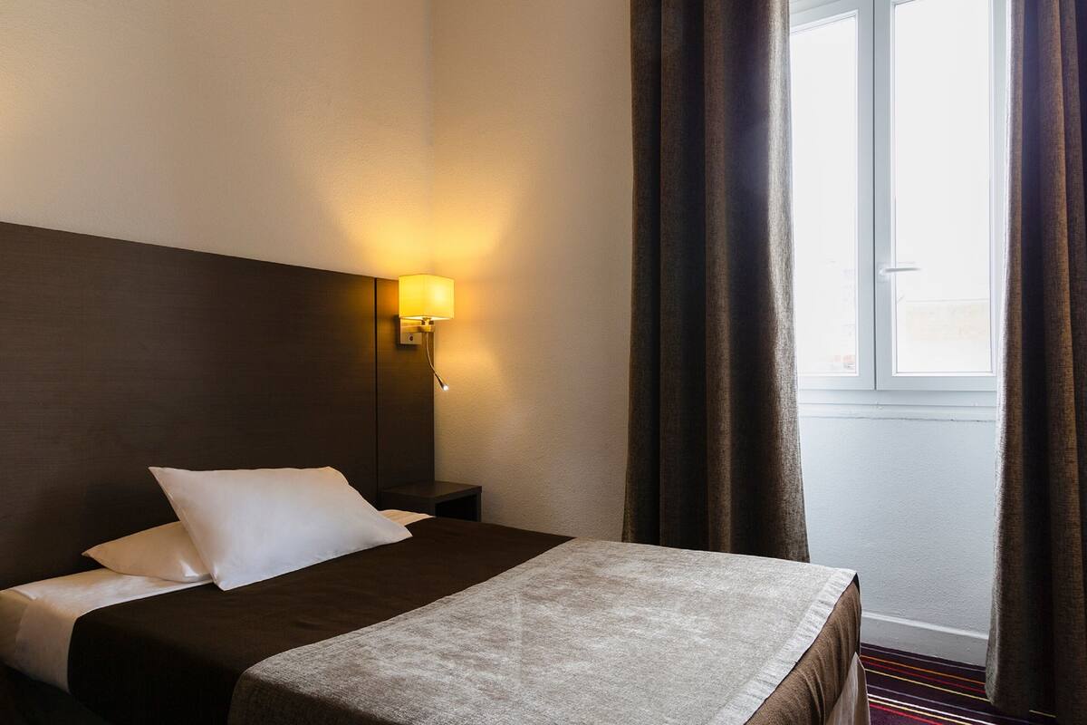 Una habitación de hotel con una cama, una mesita de noche, una lámpara y una ventana con cortinas.