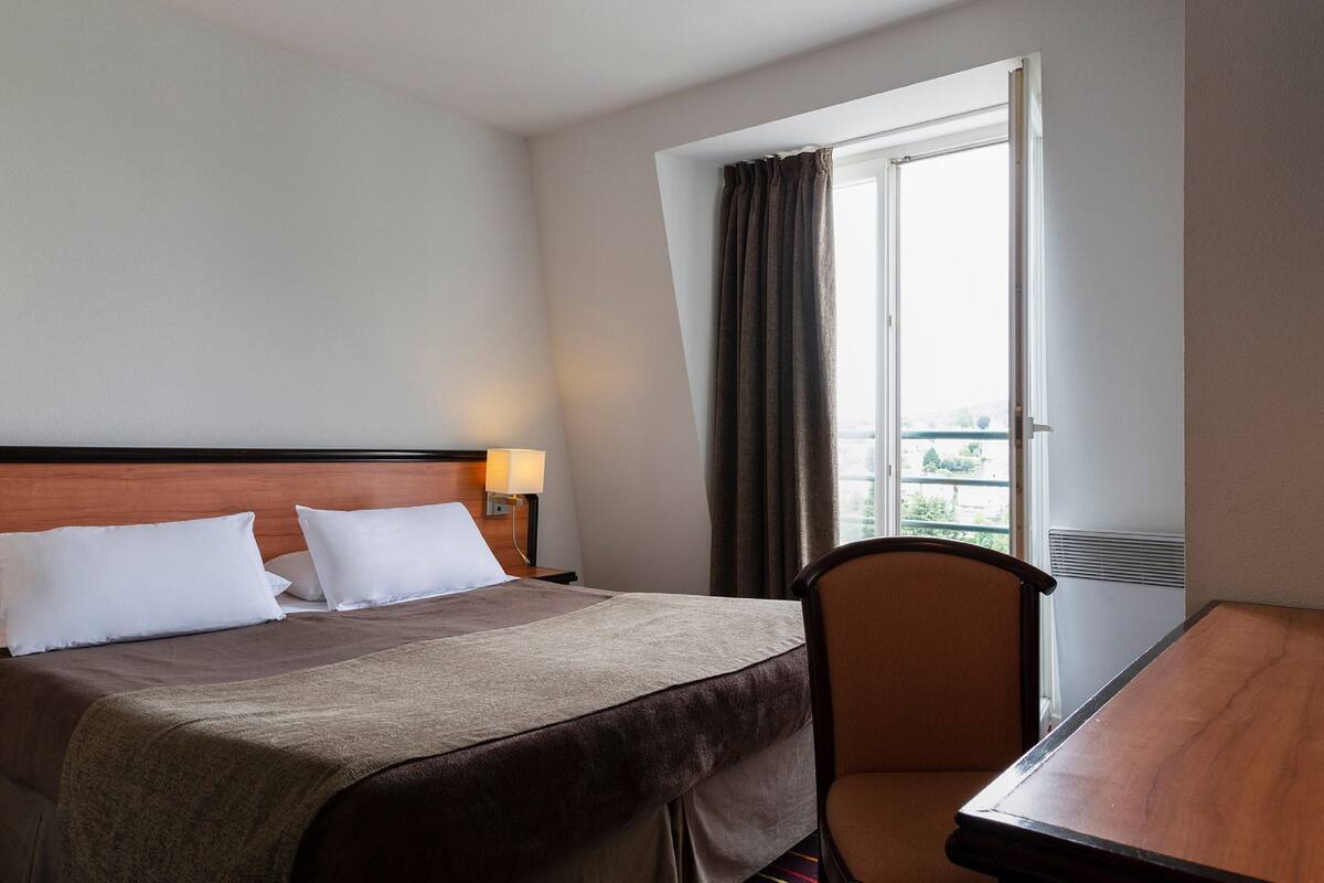 Habitación de hotel con cama, escritorio, silla, ventana con cortinas y una lámpara fijada a la pared.
