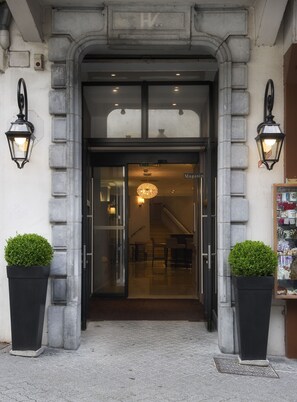 Step into a classic establishment with a stone archway and elegant entrance, flanked by lush greenery and a modern chandelier.