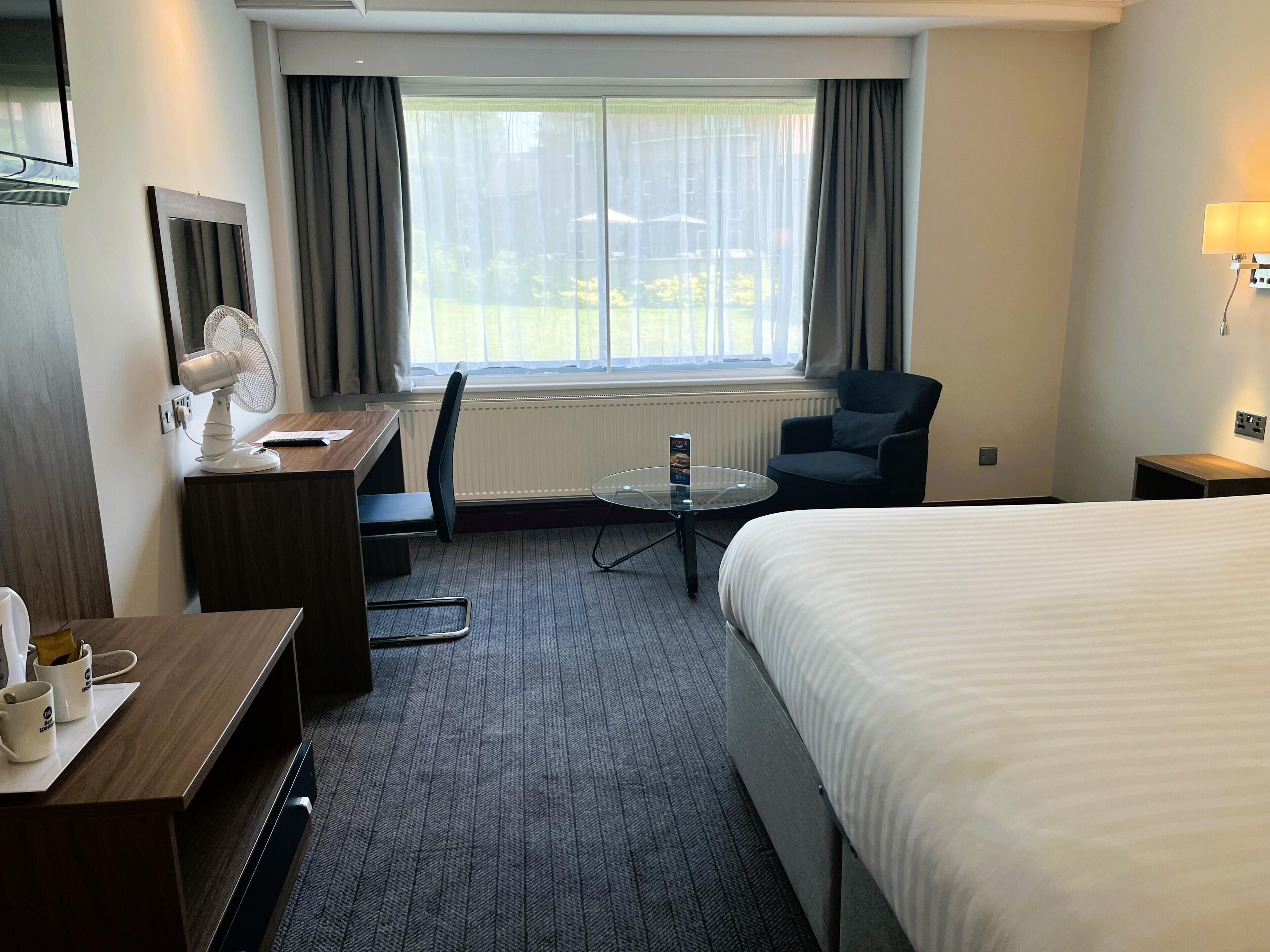 A hotel room with a bed, a desk with a chair, a small table, a fan, and a window with curtains.