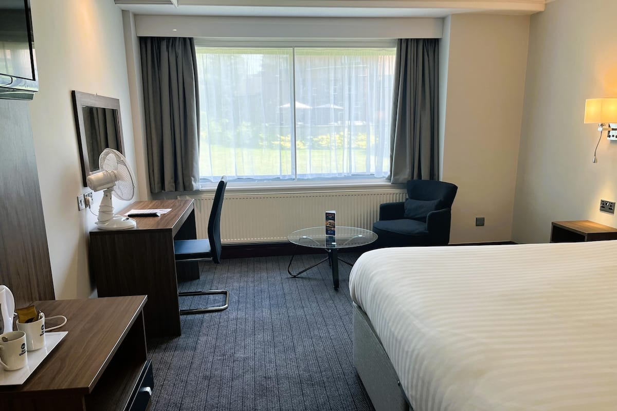 A hotel room with a bed, a desk with a chair, a small table, a fan, and a window with curtains.