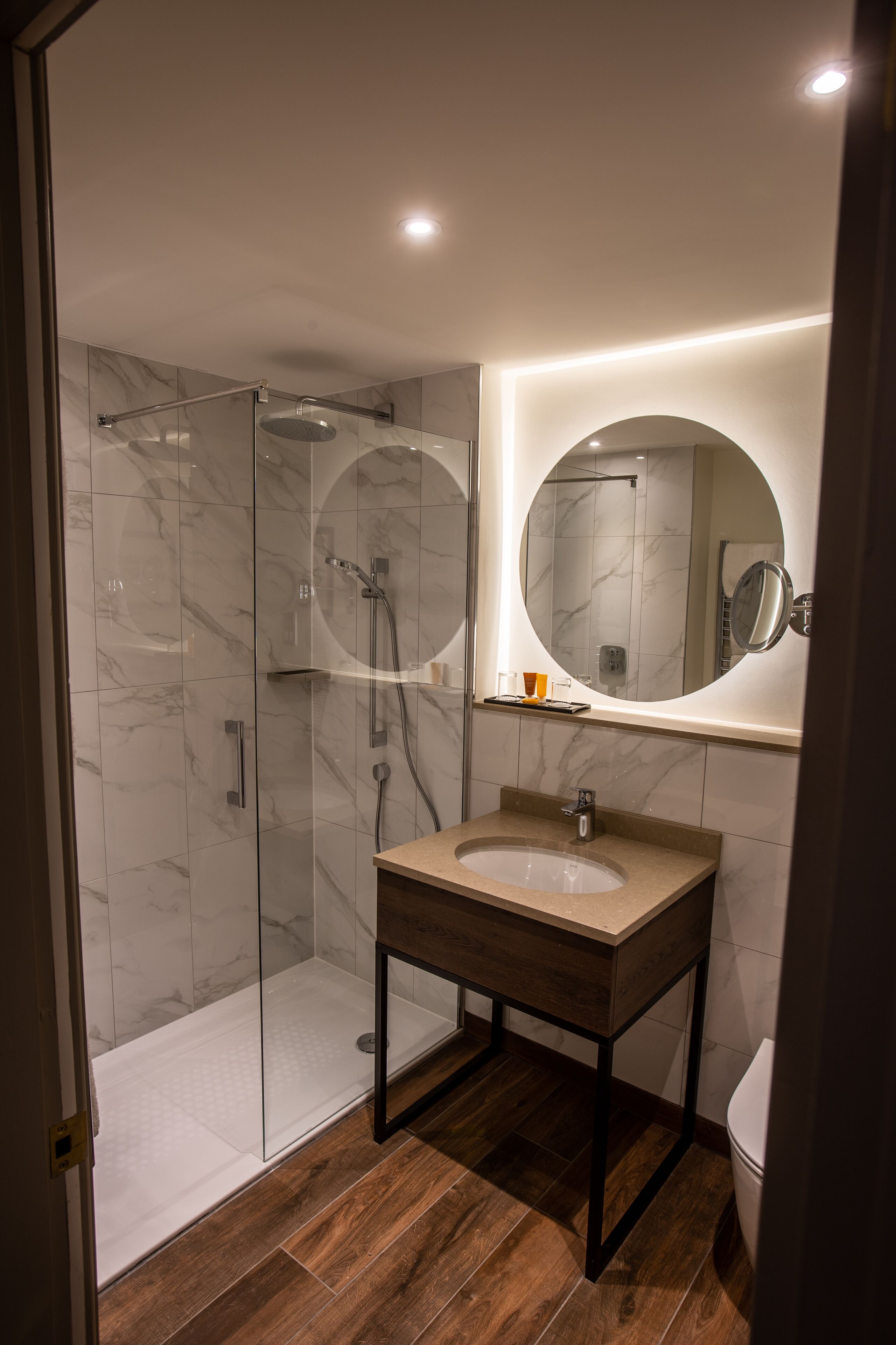 A modern bathroom with a glass-enclosed shower, a wooden vanity, and a round mirror.