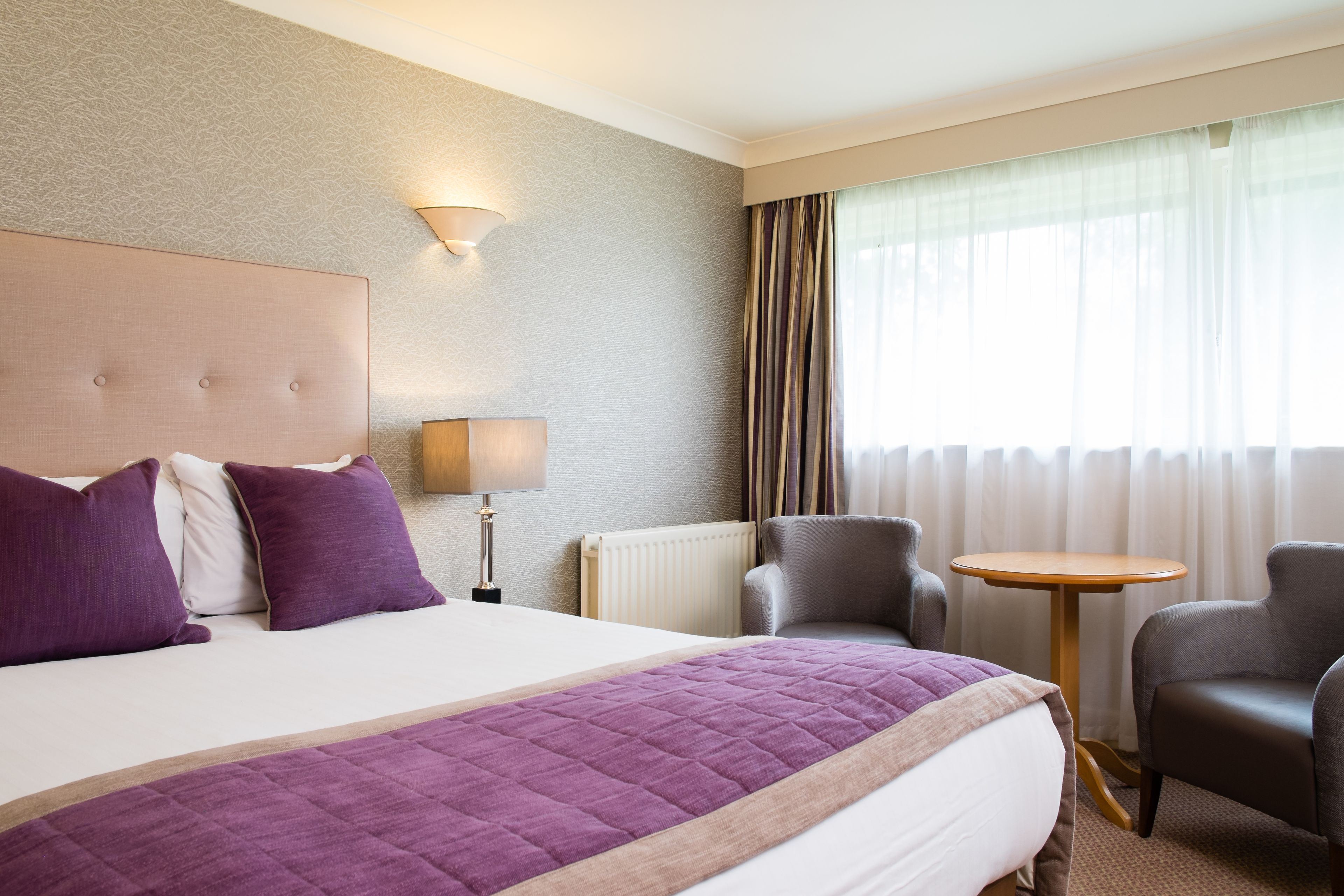 Una habitación de hotel con una cama grande, una mesita de noche, una lámpara, una silla y una pequeña mesa.
