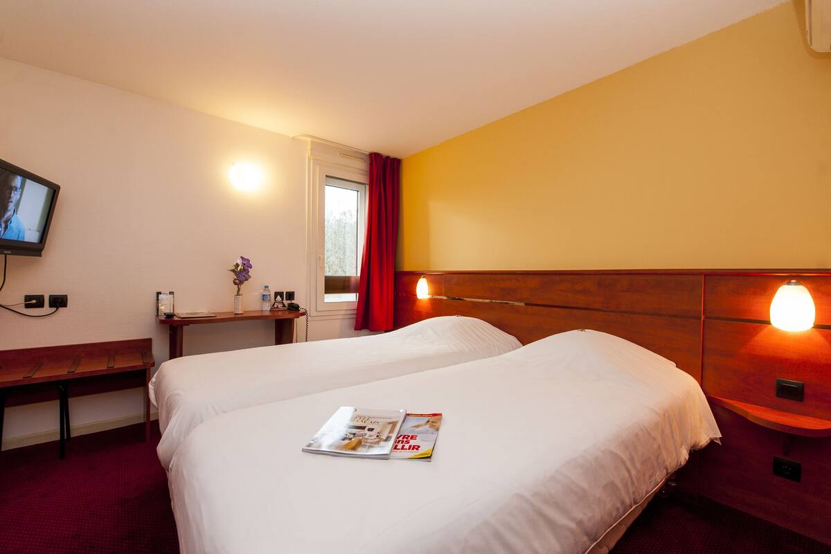 Een hotelkamer met twee bedden, een televisie, een nachtkastje met lamp en een raam met rode gordijnen.
