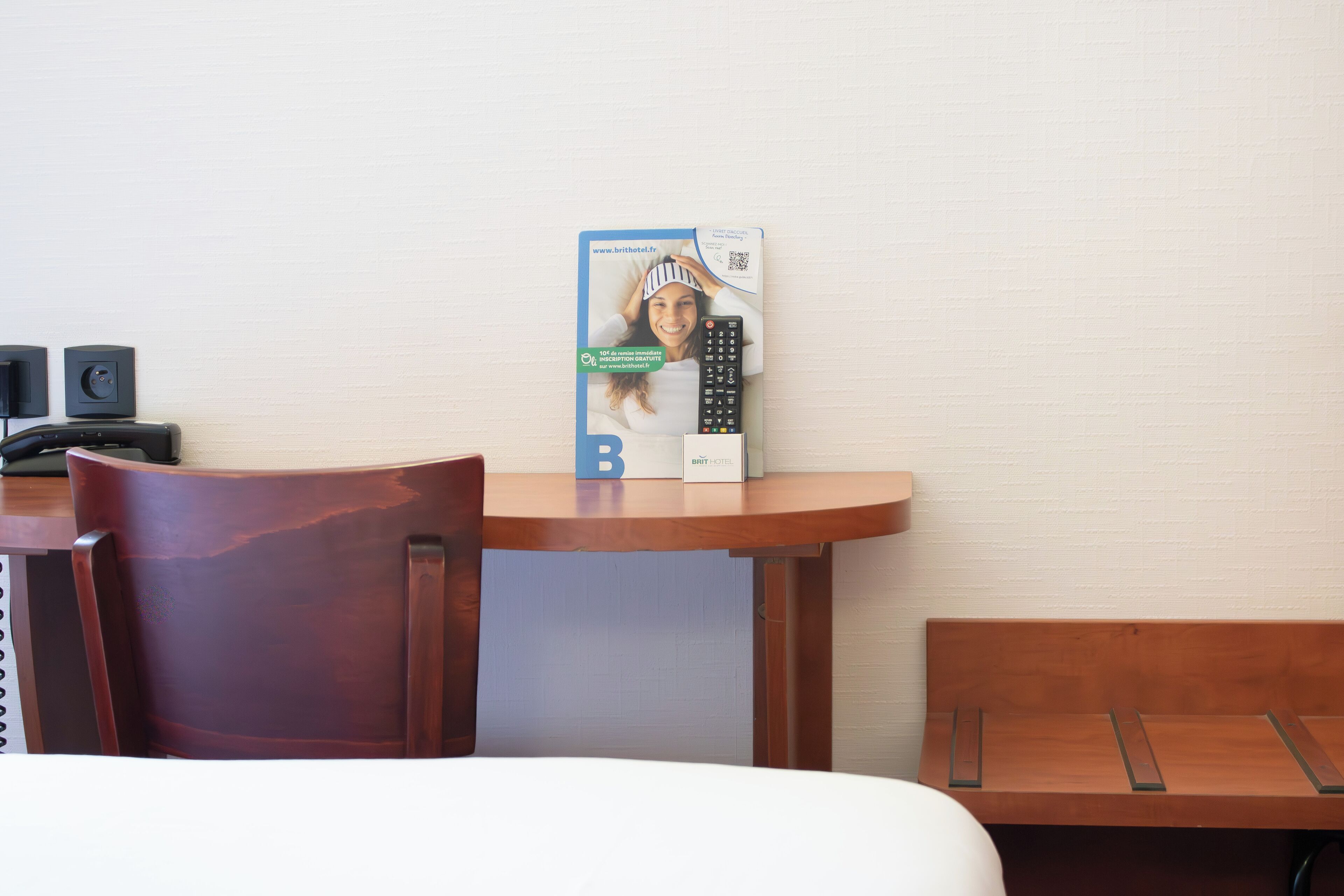 A hotel room with a wooden desk, a chair, a bed, and a reception area with a sign and a remote control.