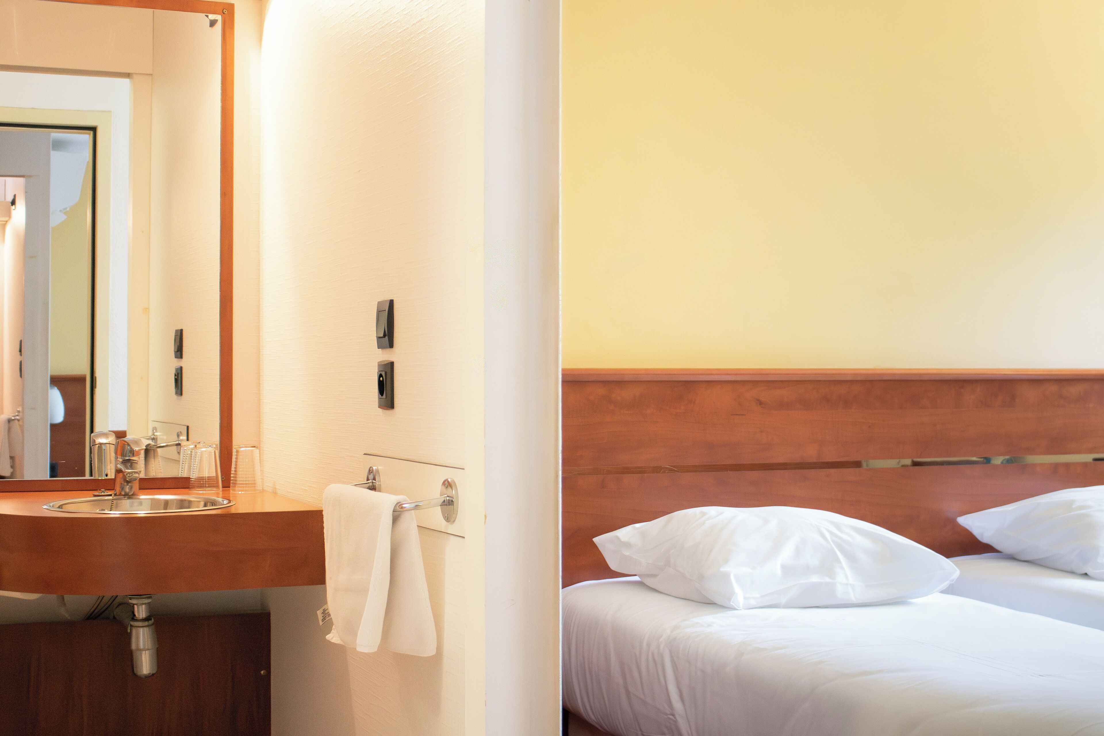 Quarto de hotel com cabeceira de madeira, cama branca e banheiro com pia e espelho.