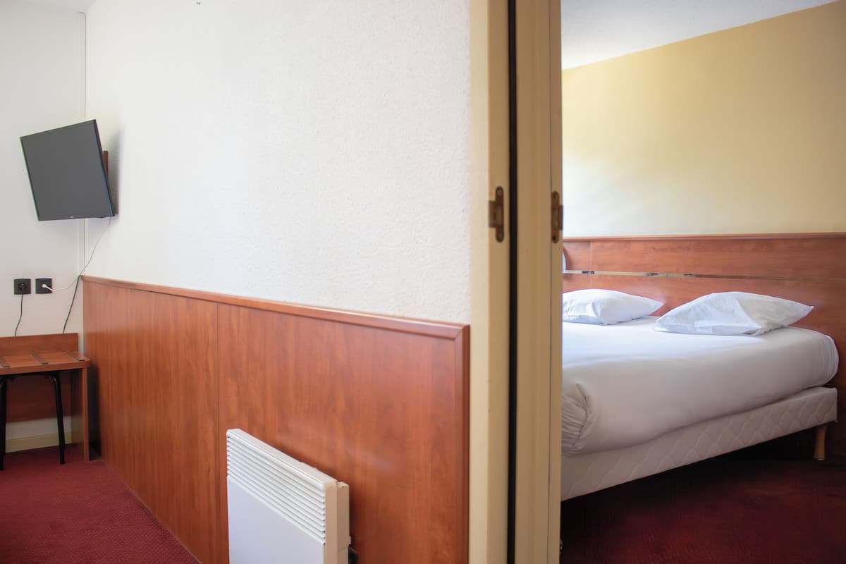 Een hotelkamer met een houten hoofdeinde, een bed met wit beddengoed, een klein houten tafeltje en een aan de muur bevestigde televisie.