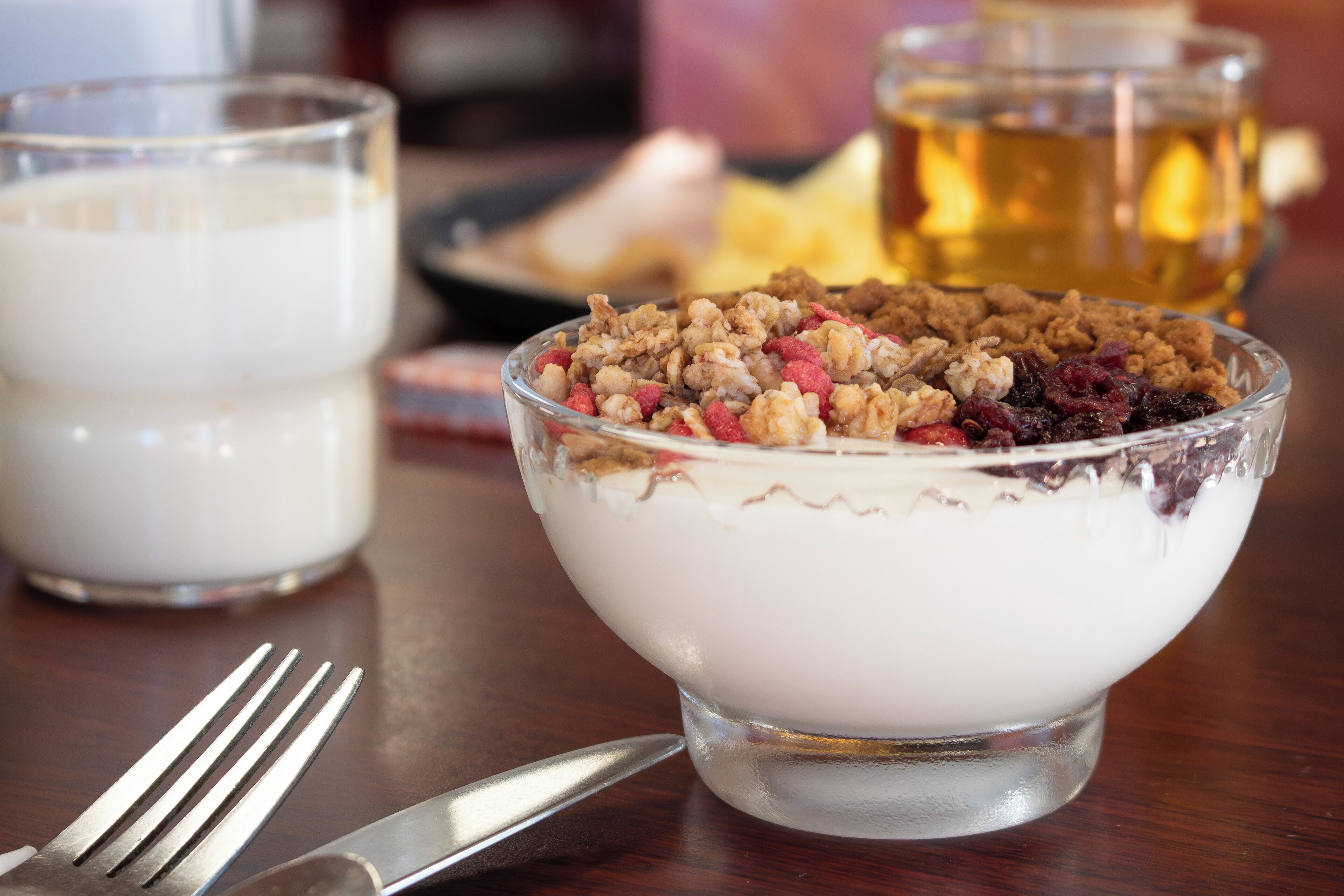 A dessert bowl with a creamy base and a topping of granola and berries, accompanied by a glass of milk and a cup of tea on a wooden table.