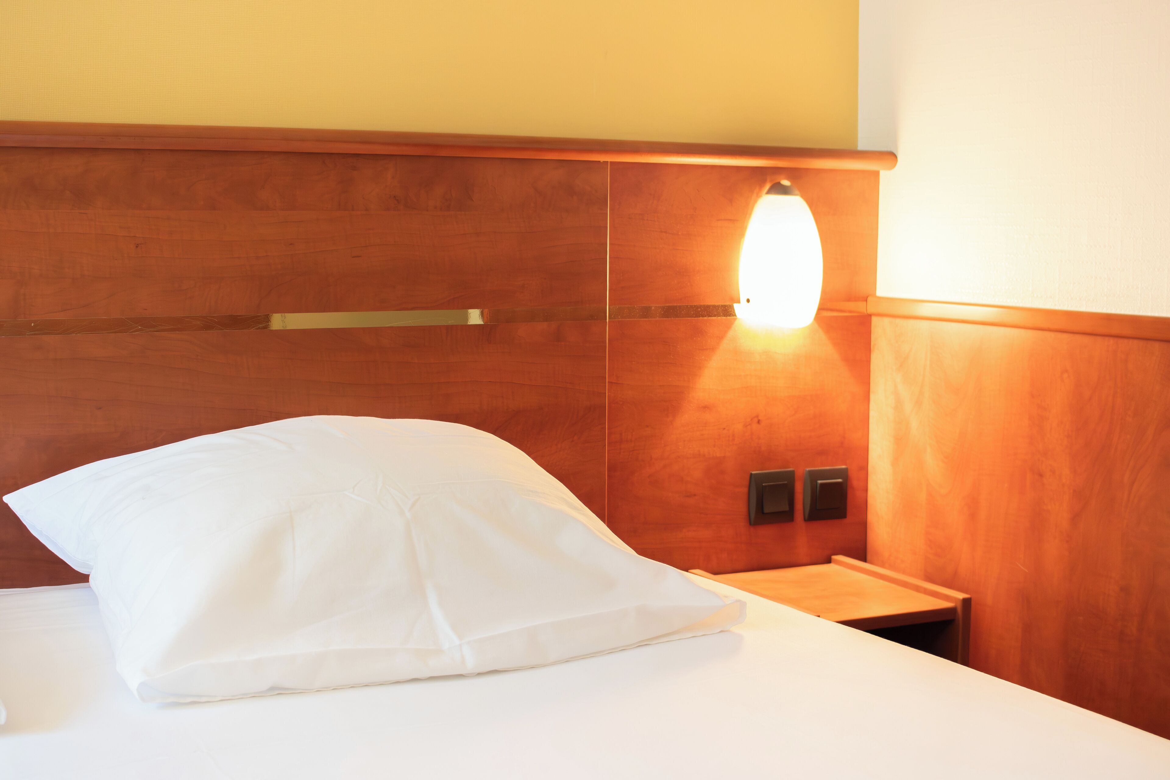 Una camera d'albergo con una testiera di legno, un comodino e un cuscino bianco.