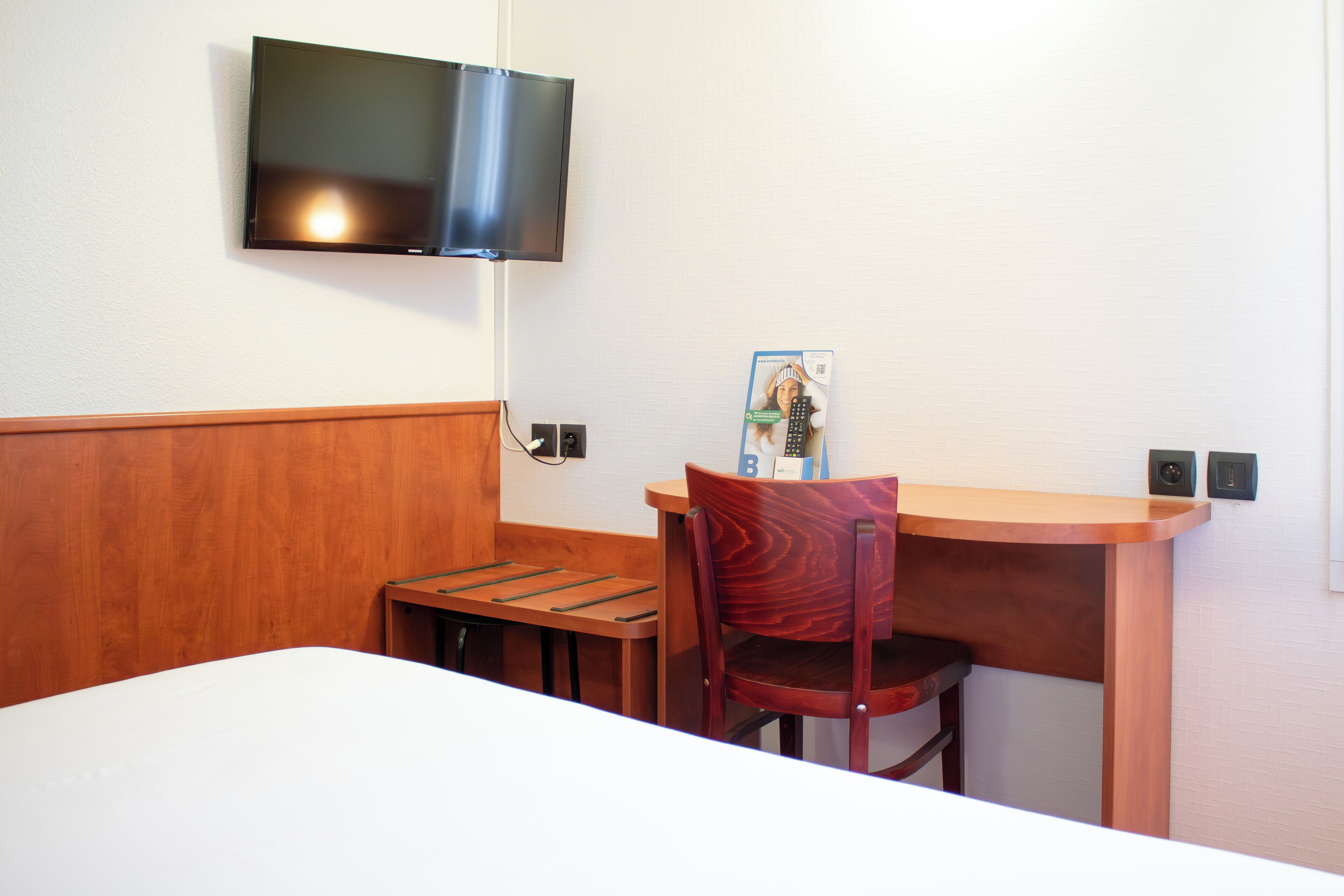 A hotel room with a bed, a desk, a chair, a television mounted on the wall, and a small table.