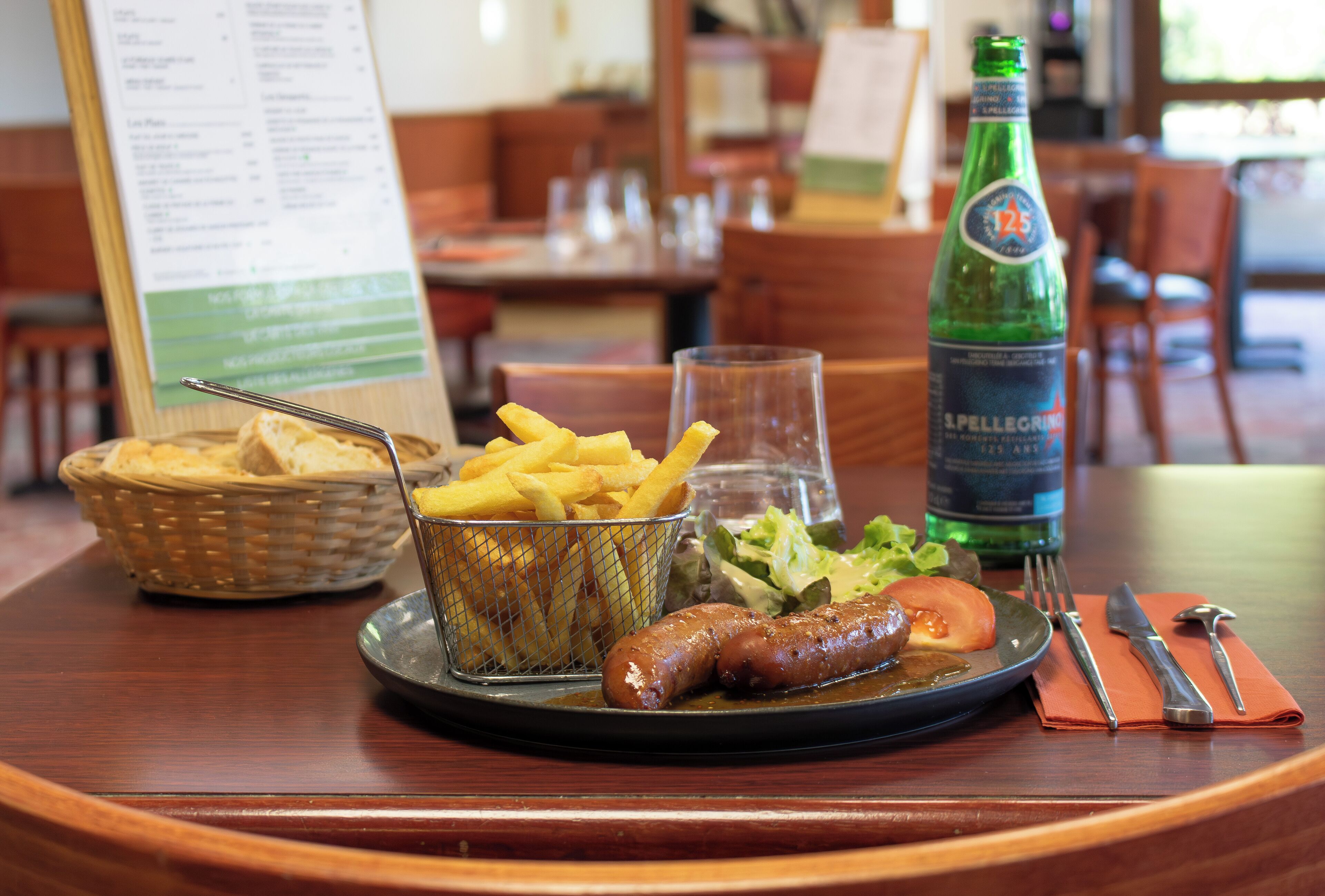 A dining table with a basket of bread, a plate with sausages and fries, a bottle of Pellegrino water, and a menu.