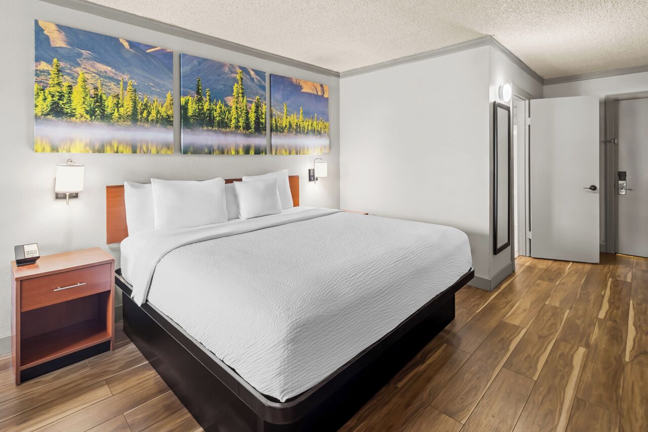 A hotel room with a large bed, a nightstand, a wall-mounted lamp, and a door with a keycard lock.