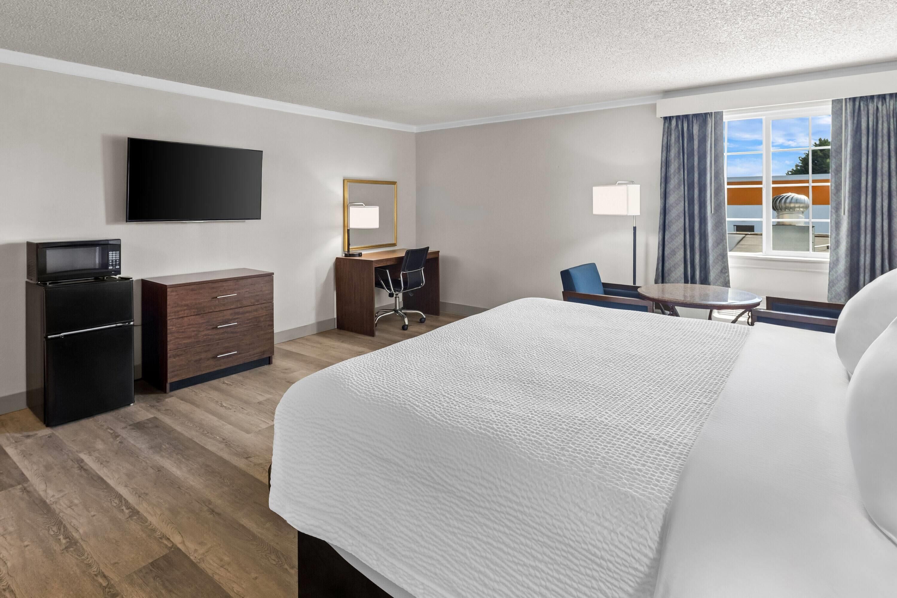 A hotel room with a bed, a desk with a chair, a television, a microwave, and a window with curtains.