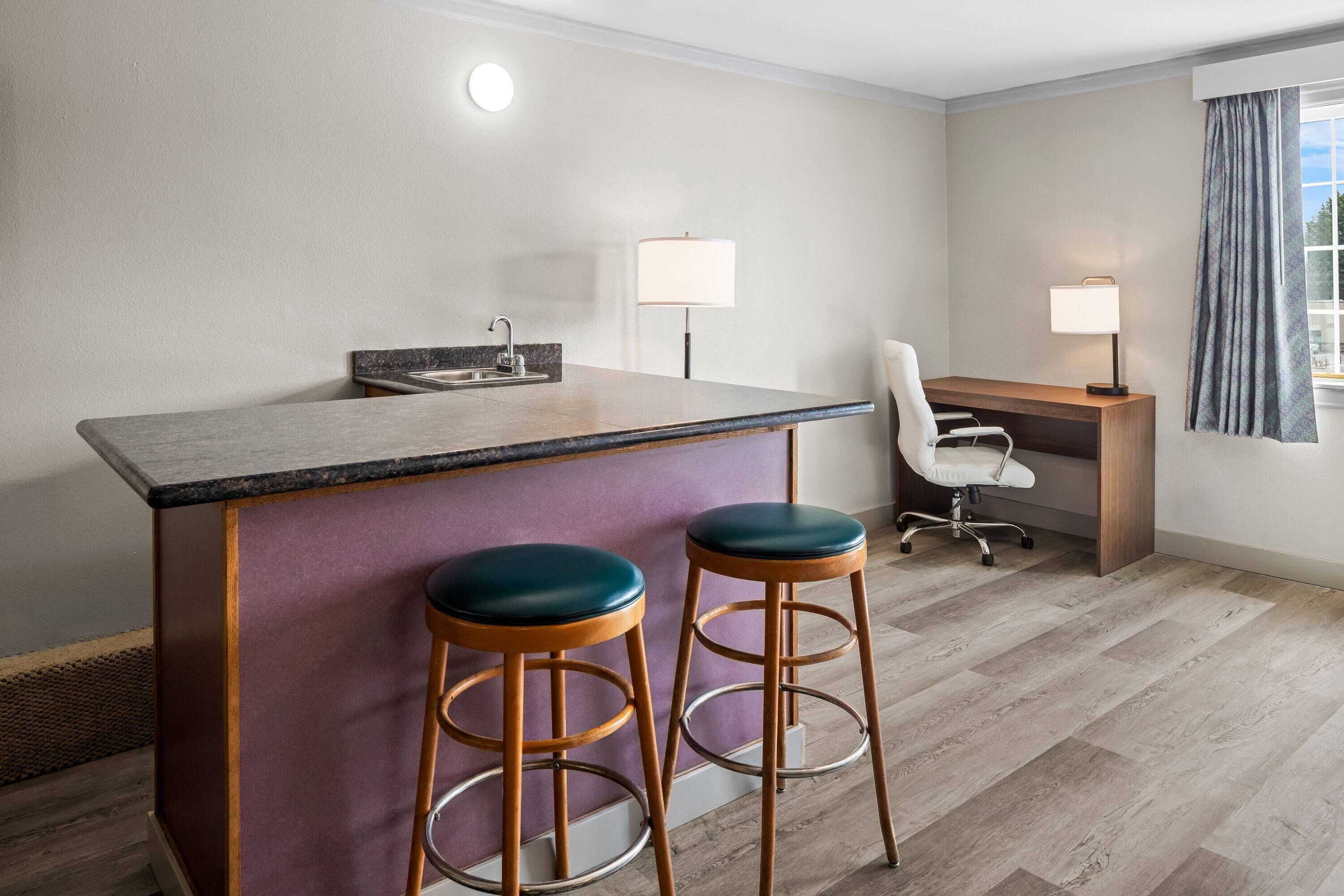 A modern hotel room with a kitchen area, two bar stools, a desk, and a chair.