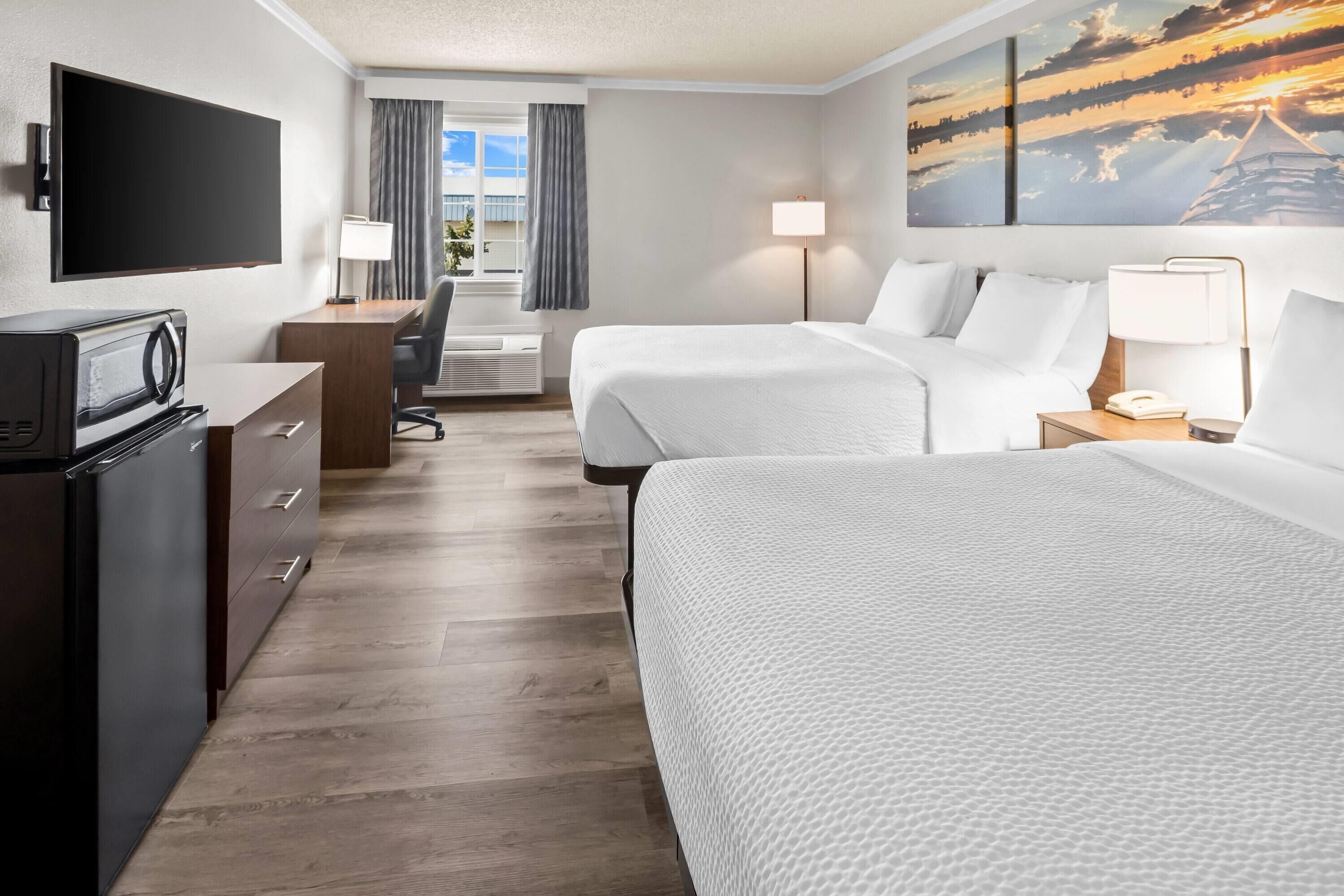 A hotel room with two beds, a TV, a desk, a chair, a lamp, and a painting.