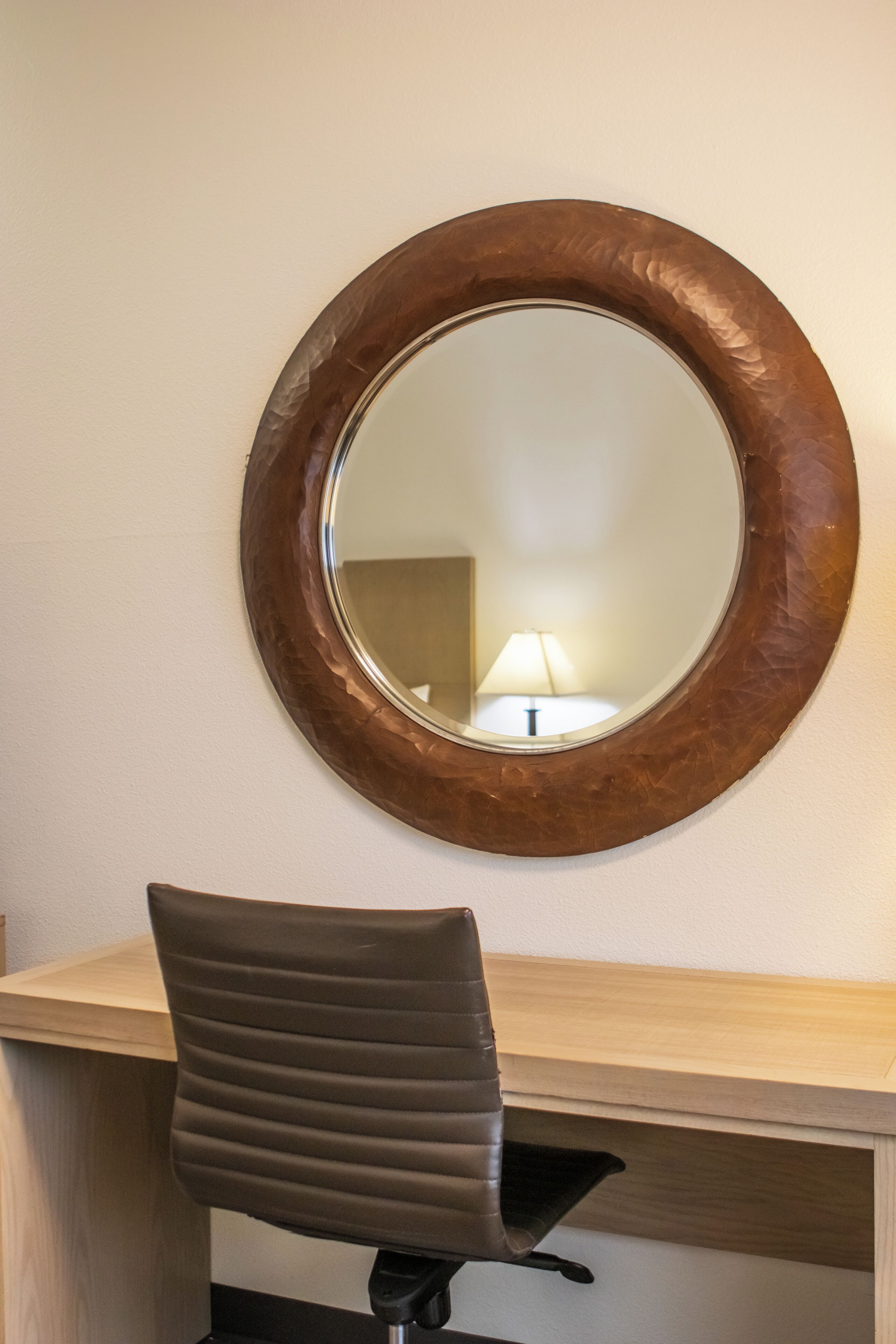 Un espejo redondo con marco de madera en una pared, un escritorio con una silla y una lámpara en el reflejo.