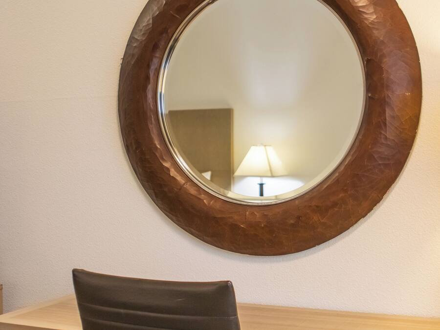 A round mirror with a wooden frame on a wall, a desk with a chair, and a lamp in the reflection.