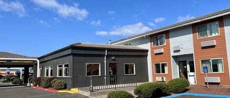 A building with a grey facade and a brown wooden section, featuring a gated entrance and a parking lot with marked spaces.