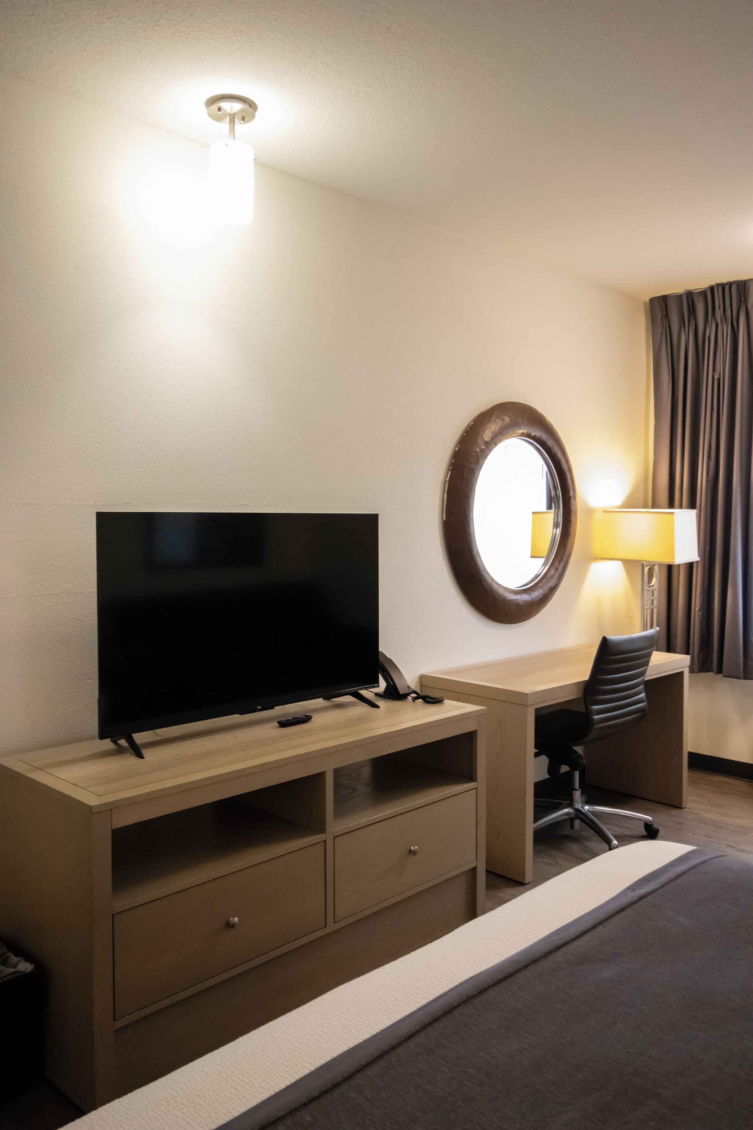 Ein Hotelzimmer mit Flachbildfernseher, Schreibtisch mit Lampe, Stuhl, Spiegel und Bett.